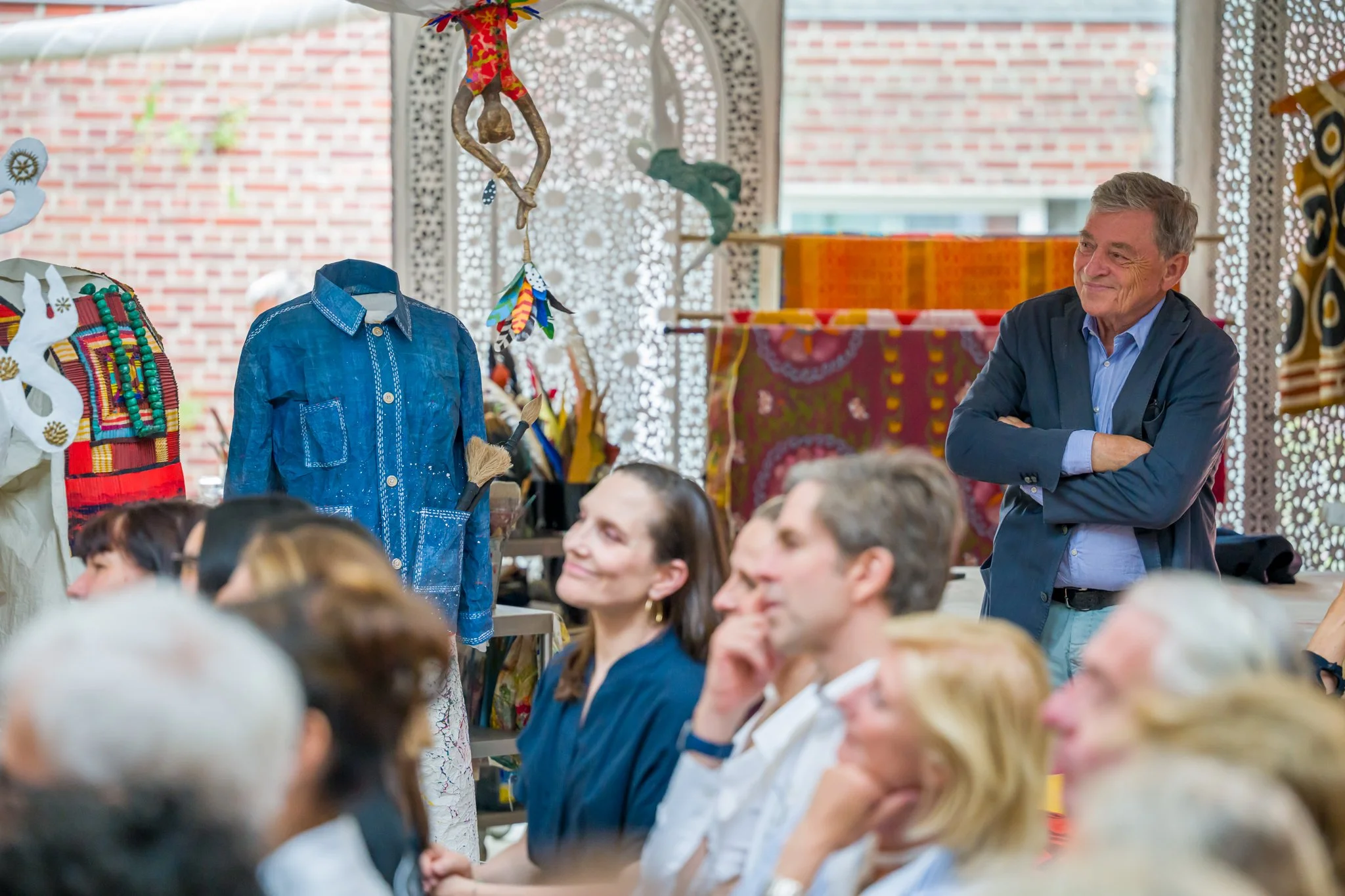 Une réunion ou une présentation avec un groupe de personnes assises et un homme debout qui observe, dans un décor coloré avec des vêtements et objets artisanaux exposés.