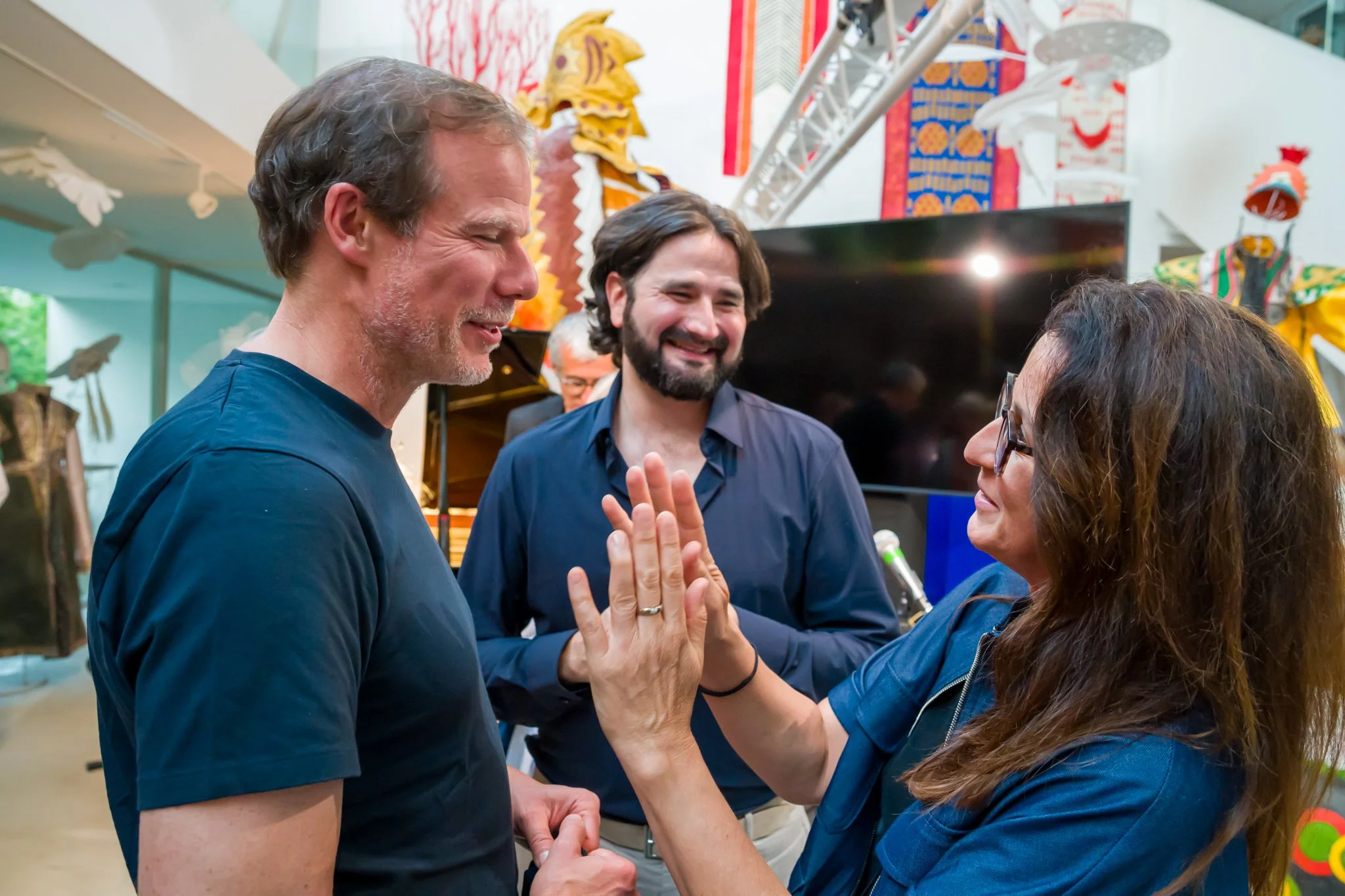 Trois personnes souriantes discutant et se saluant dans un lieu décoré avec des objets colorés et des œuvres d'art. La femme fait un geste de bienvenue avec la main, tandis que deux hommes la regardent avec amusement et amitié.