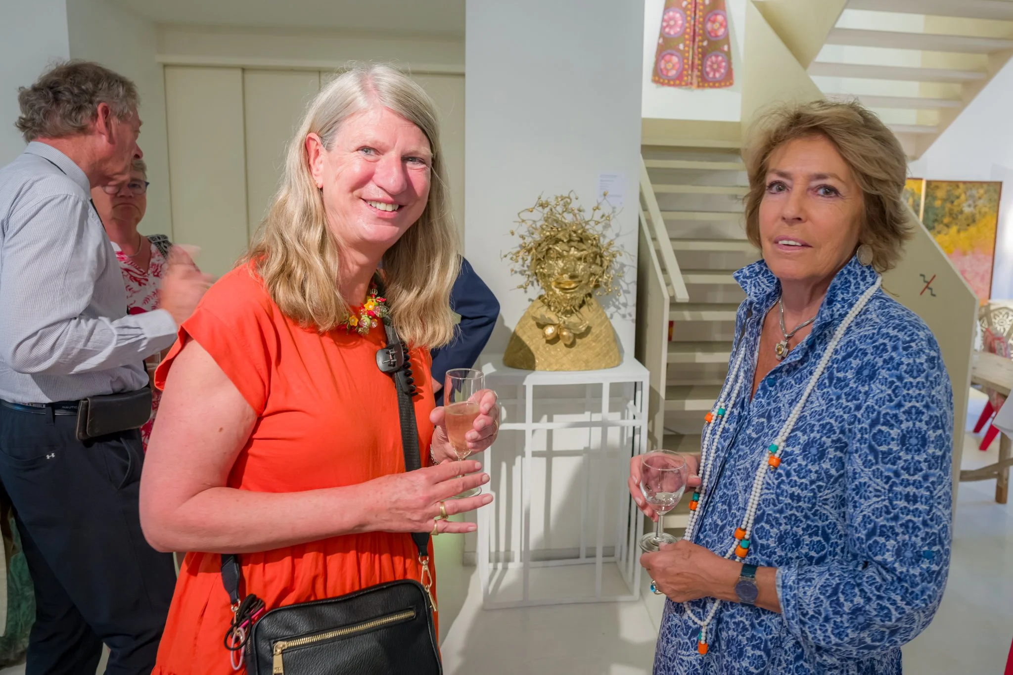 Deux femmes se tenant un verre à la main dans une galerie d'art, souriantes, avec d'autres personnes en arrière-plan et une sculpture dorée derrière elles.