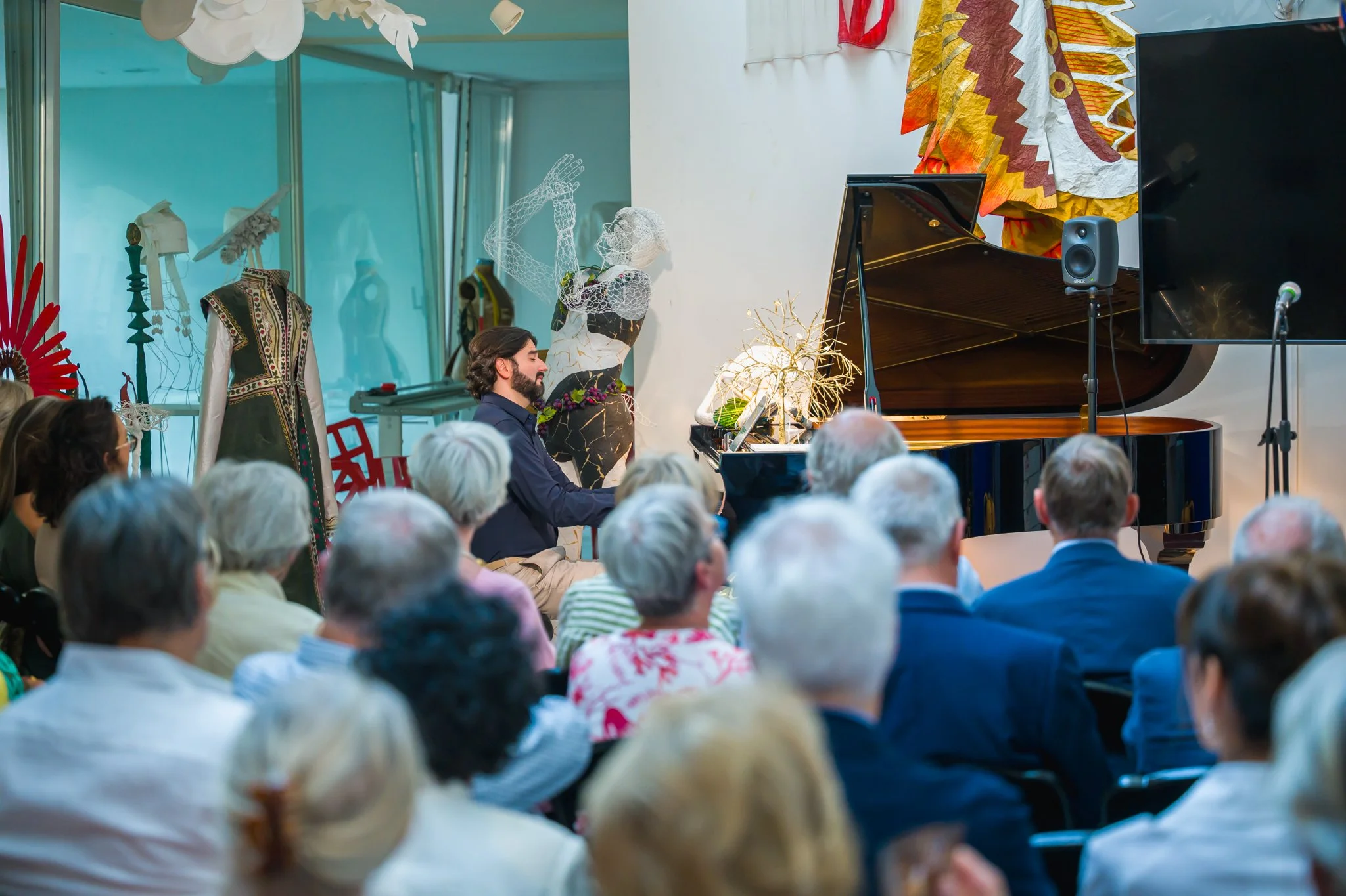 Un homme joue du piano lors d'un concert devant un public d'adultes âgés, dans une salle décorée avec des œuvres d'art et des sculptures.