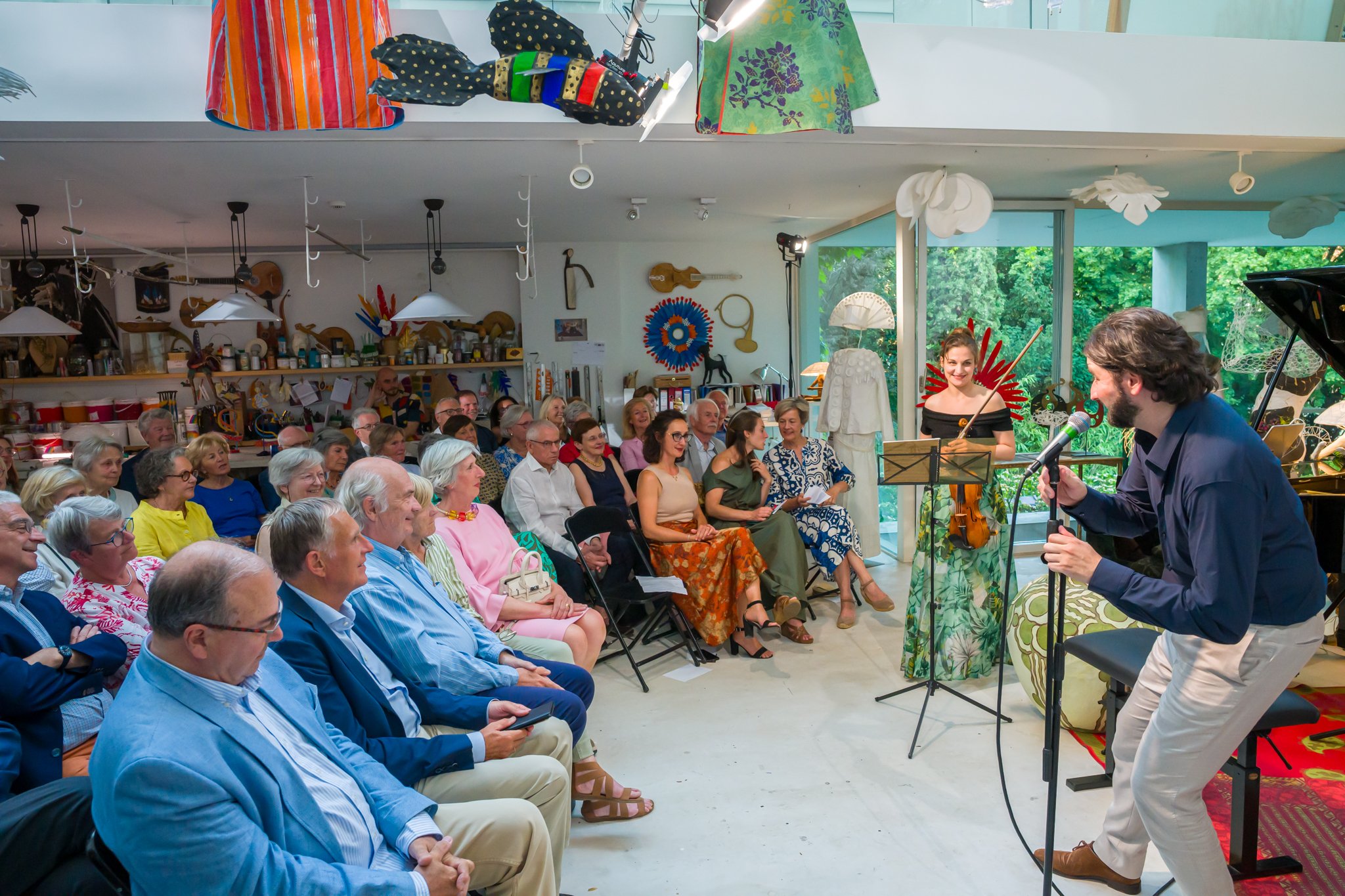 Concert de musique avec un homme chantant et un quatuor à cordes devant un public dans une salle d'art ou artisanat, avec des objets décoratifs suspendus au plafond et des œuvres d'art au mur.