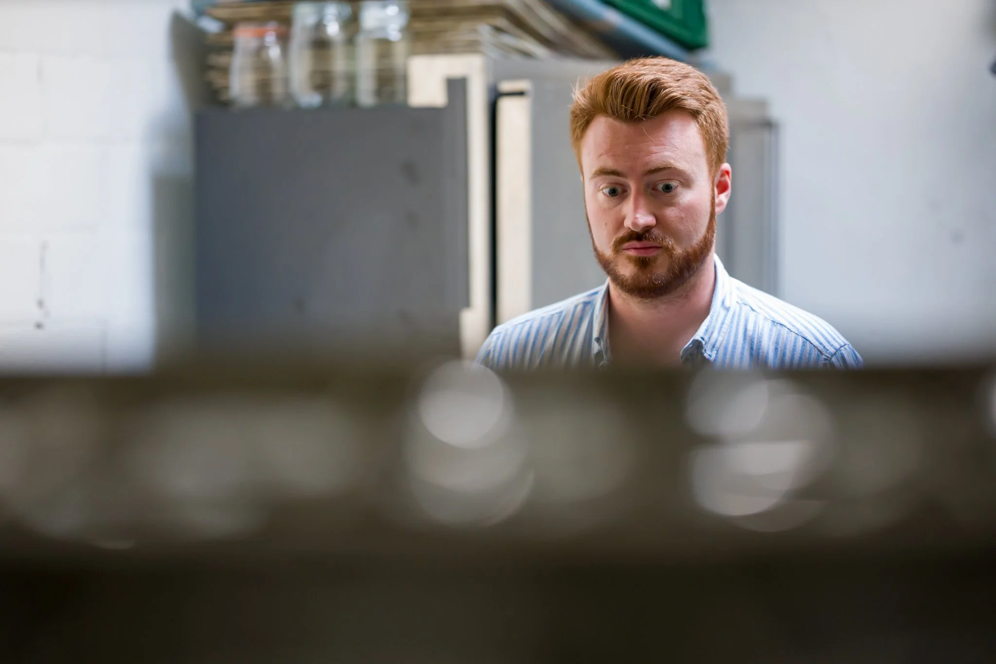 Un homme aux cheveux roux et barbe rousse, portant une chemise à rayures blanches et bleues, regarde devant lui avec une expression surprise ou curieuse. L'arrière-plan montre une cuisine ou un espace de travail avec des étagères et des contenants.