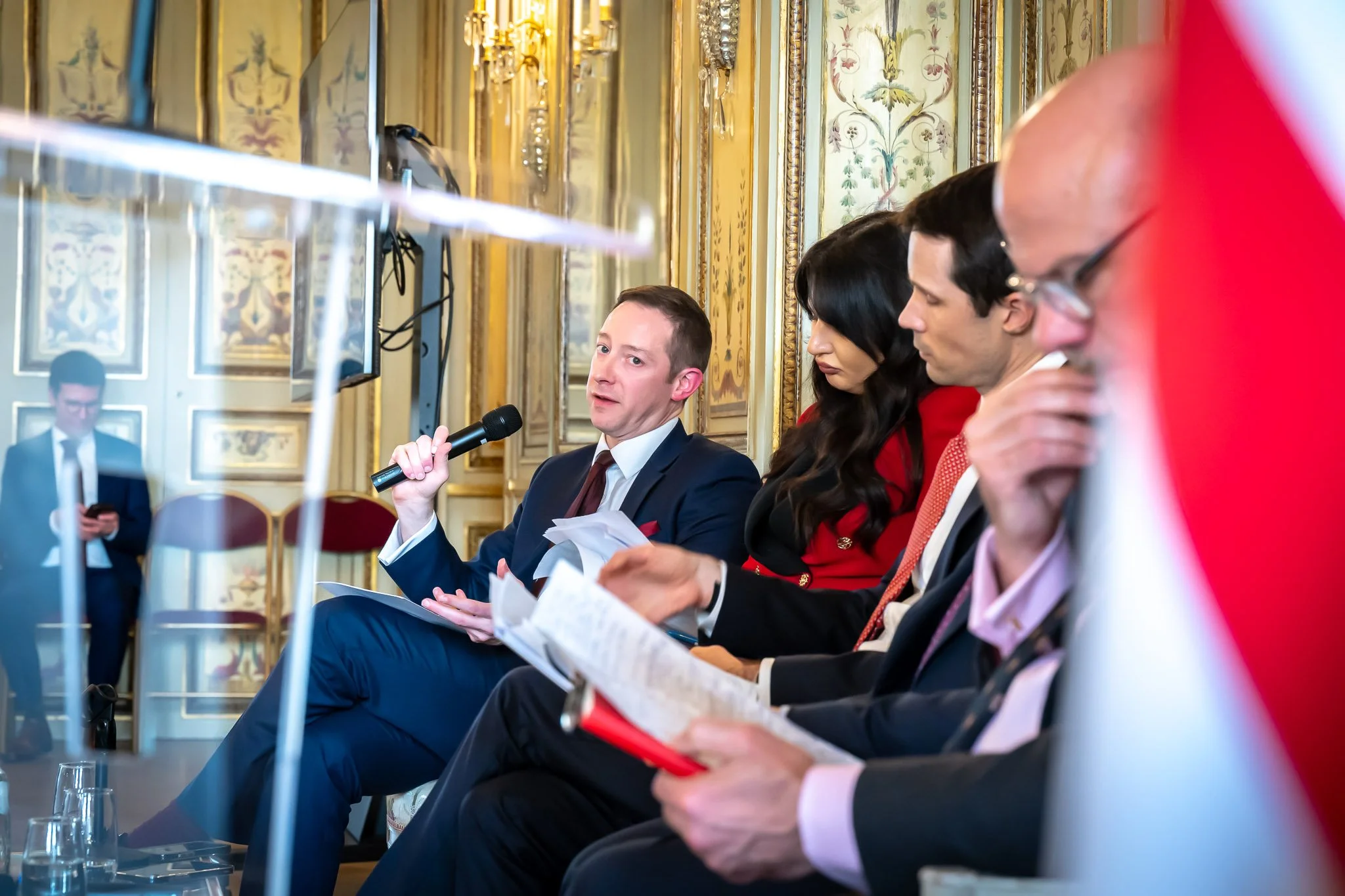 A group of professionally dressed people are attending a conference in an elegant room. One of them is speaking into a microphone, while the others listen and take notes. A man, in the background, is seated and using his TV.