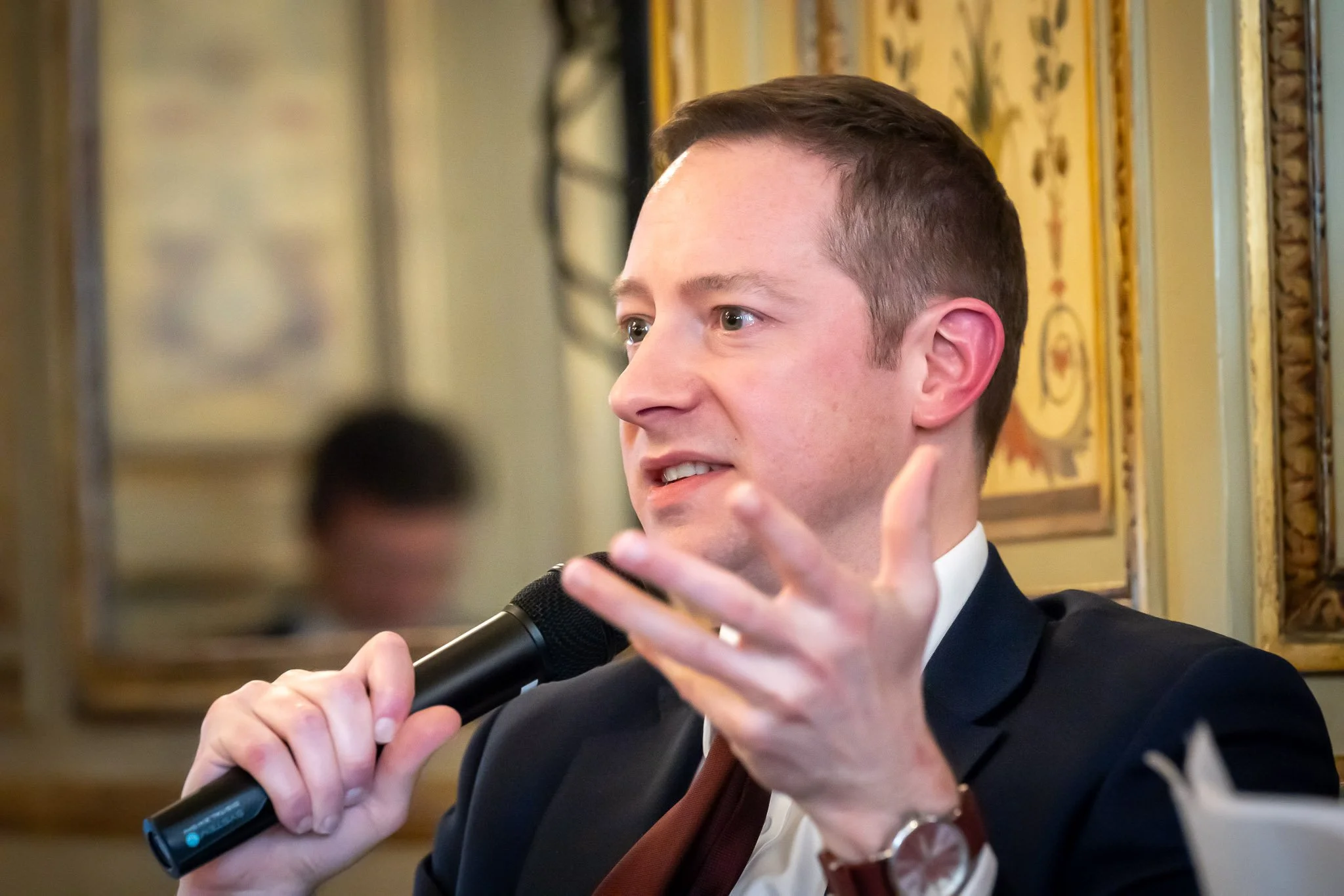 A man in a suit holding a microphone and speaking at an event in a decorative setting.