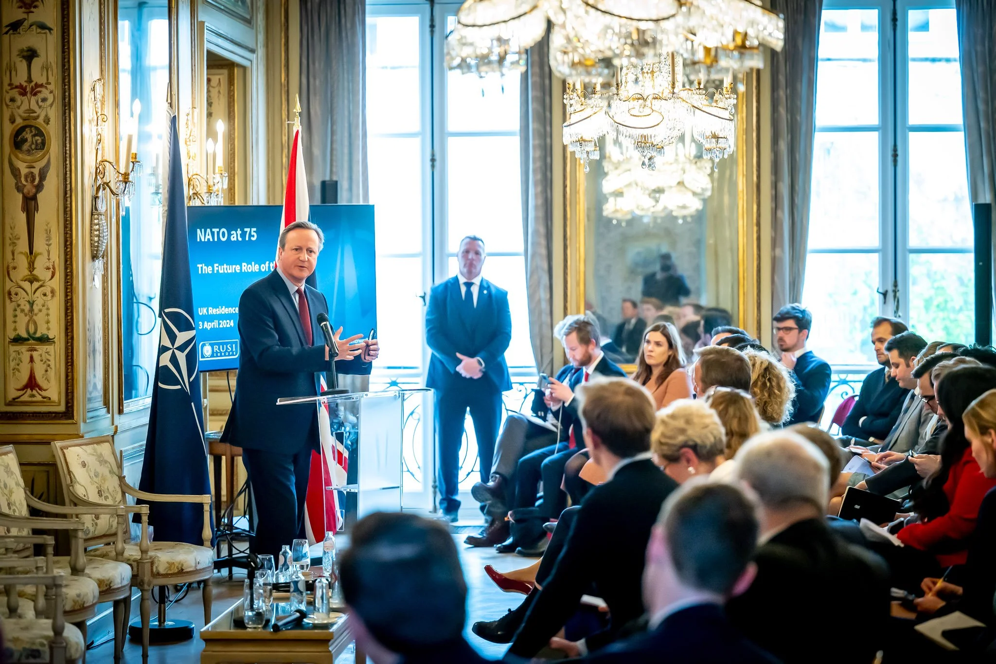 Conference on NATO at 75 with a speaker before an audience in a room decorated with chandeliers. A screen displays "NATO at 75".