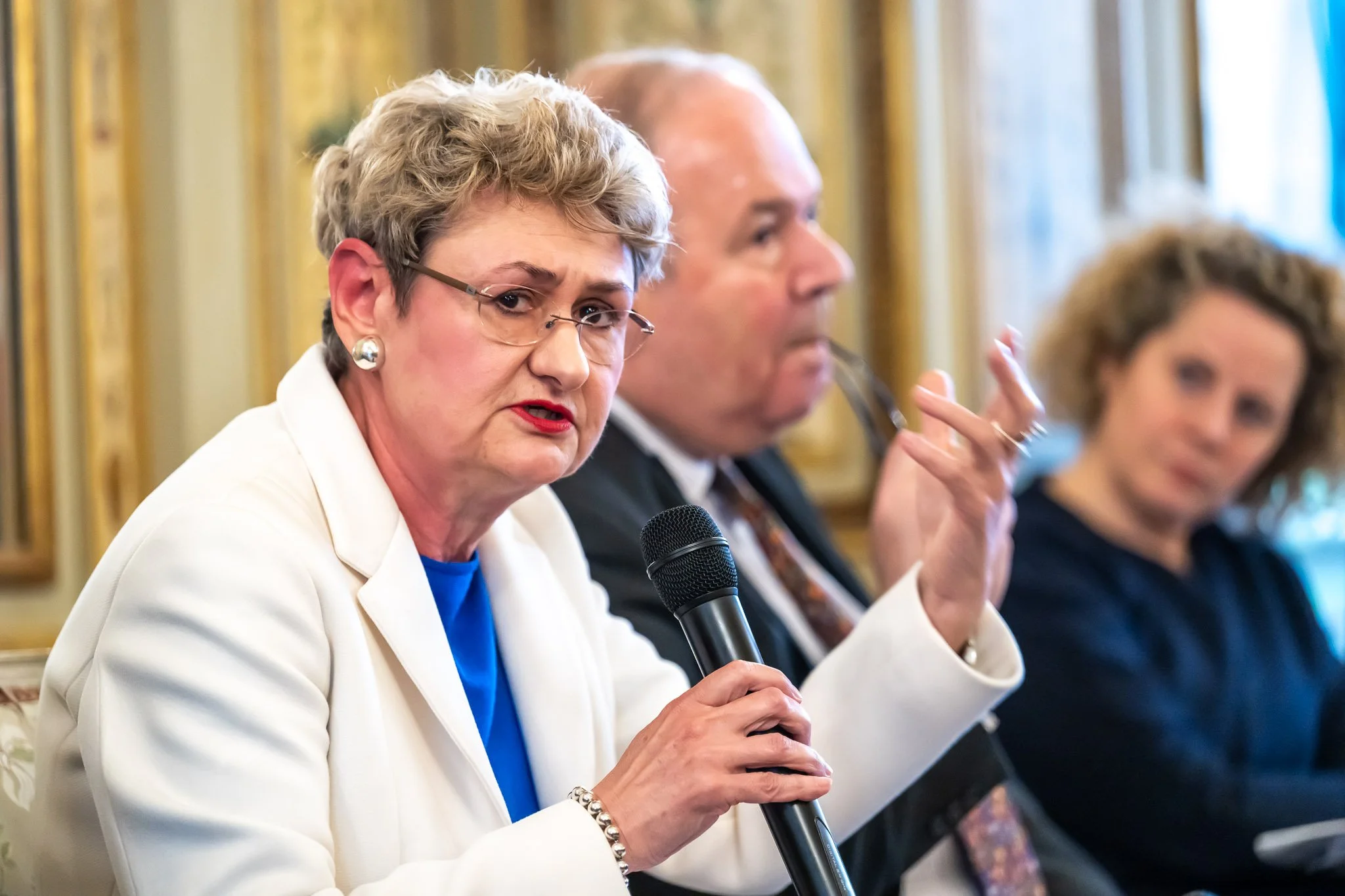 Woman with a microphone speaking at an event, with two other people blurred in the background.