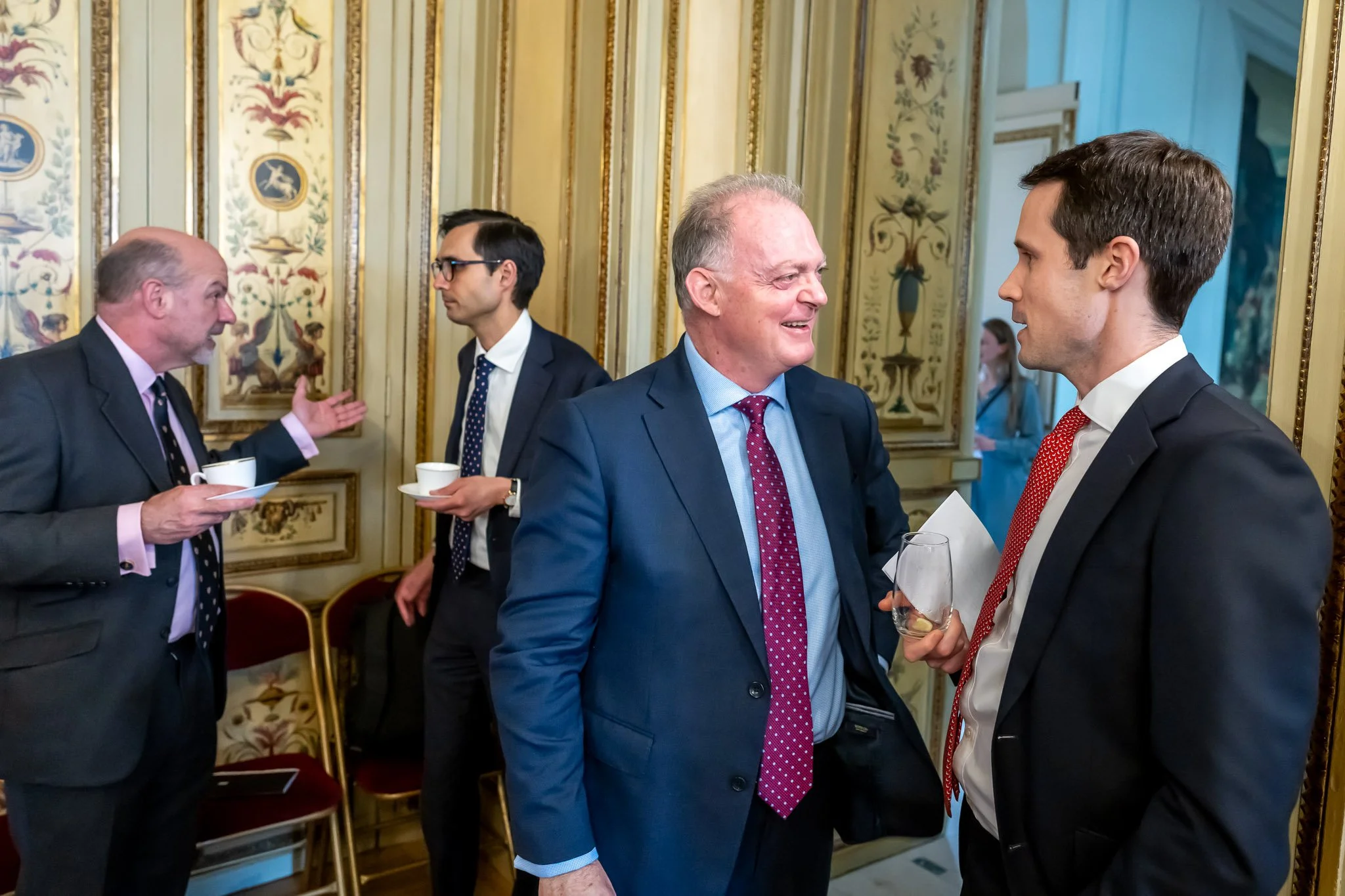 Four men in suits are chatting in an elegant room, with gilded decorative details on the walls. Two men hold cups of coffee, and another holds a glass.
