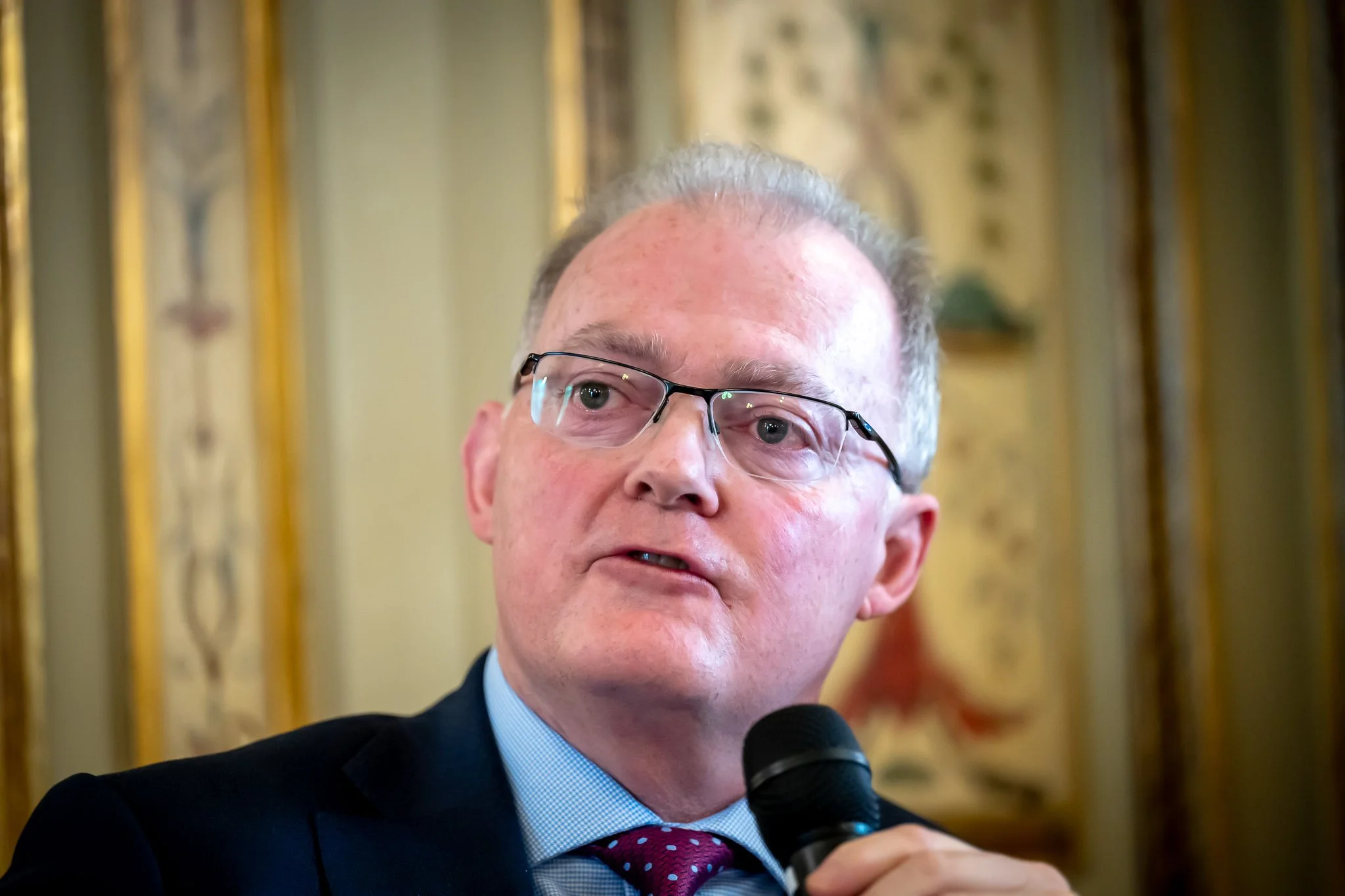 Man with glasses speaking into a microphone, decorative background.