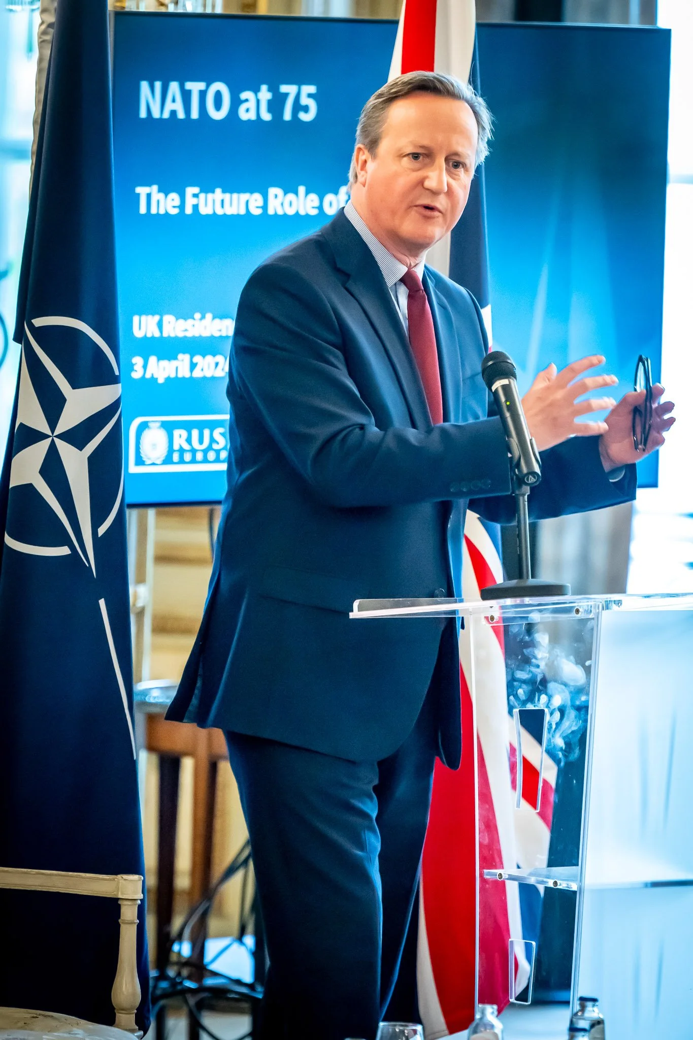 A man in a suit speaks into a microphone at an event marking 75 years of NATO, with a NATO flag in the background.