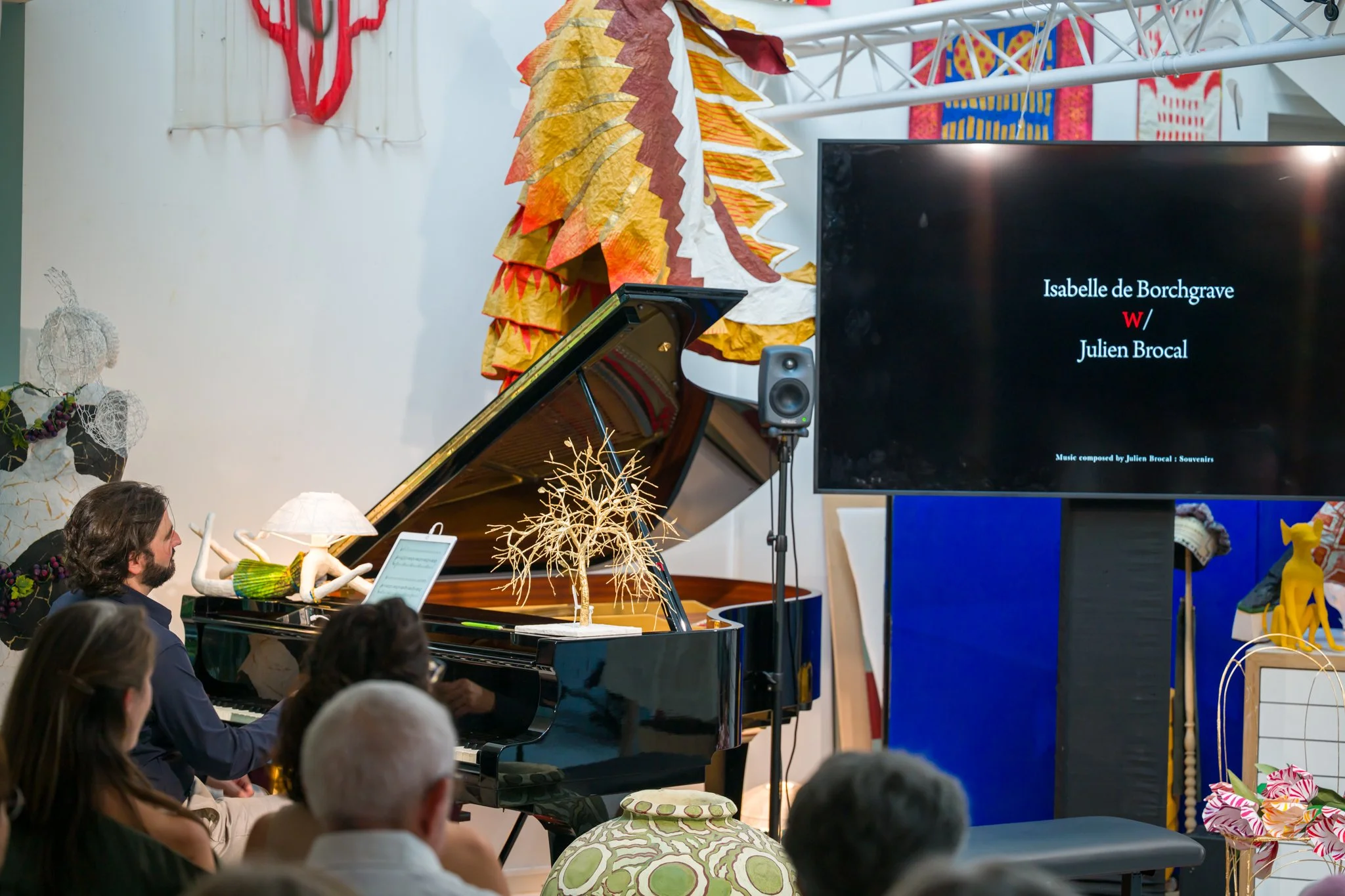 Concert ou présentation avec un piano à queue noir, un écran affichant les noms Isabelle de Borchgrave et Julien Brocal, et un petit ensemble de décorations artistiques et textiles colorés. Audience assise regardant la scène.