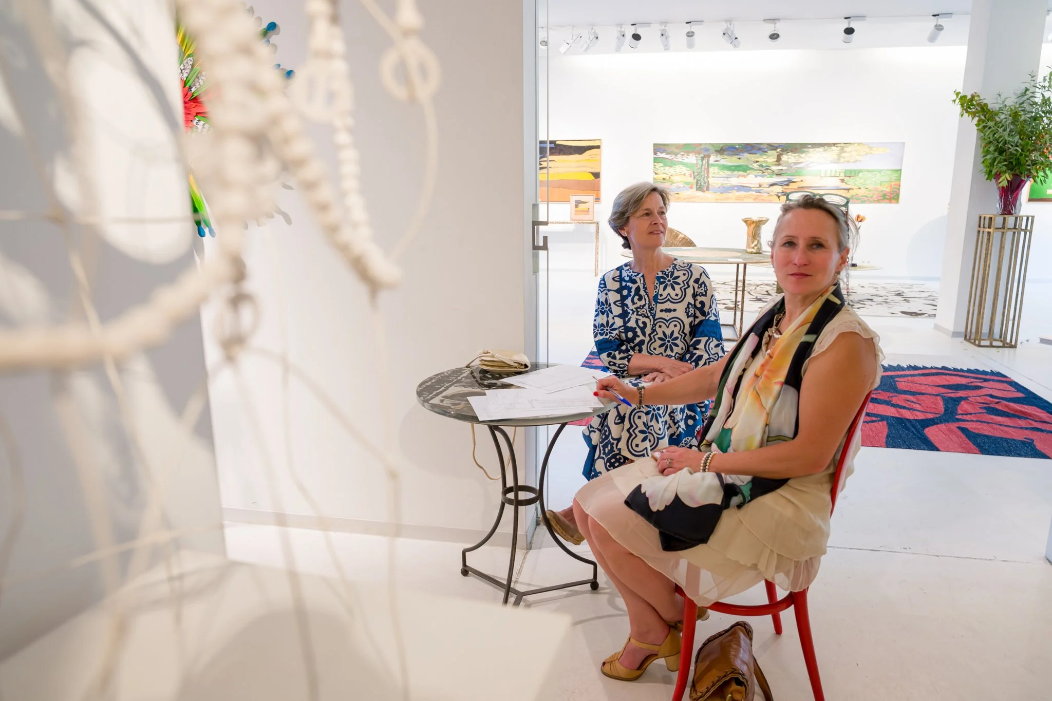 Deux femmes assises à une table dans une galerie d'art, entourées d'œuvres d'art et de sculptures, avec un tapis coloré en arrière-plan.