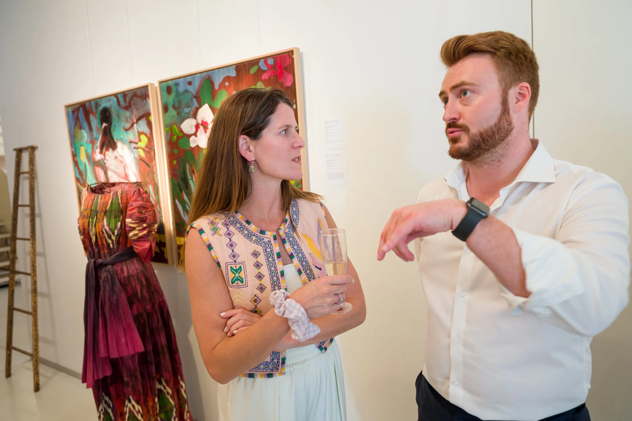 Deux personnes discutent lors d'une exposition d'art, l'une tient un verre de champagne, en arrière-plan une peinture et une sculpture en bois