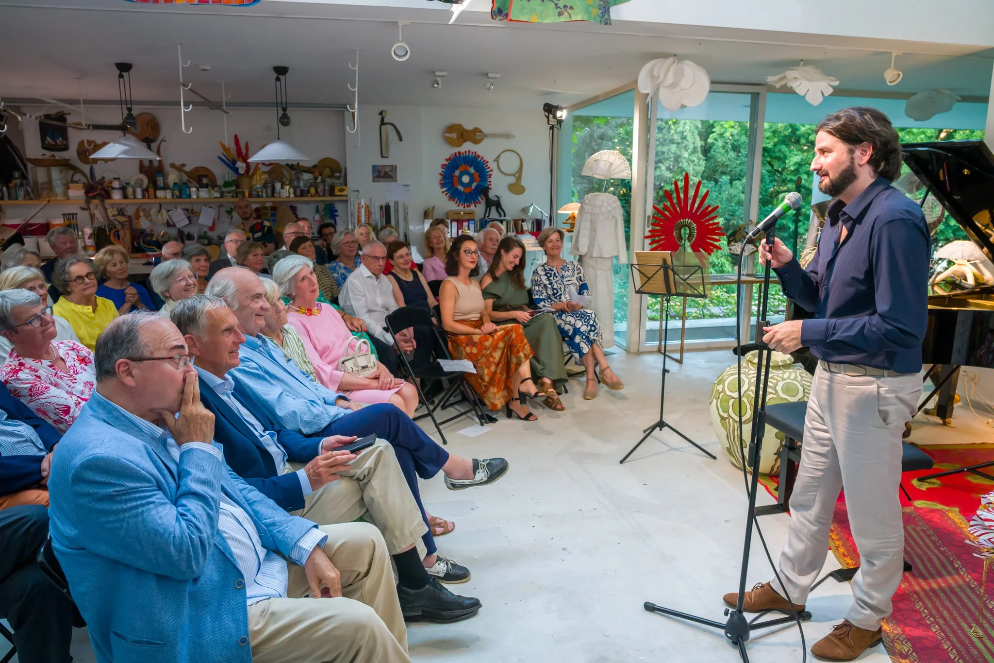 Un homme parle au micro lors d'un spectacle ou d'une présentation devant un public d'adultes dans une salle de style artistique avec des décorations colorées et une grande fenêtre donnant sur la nature.