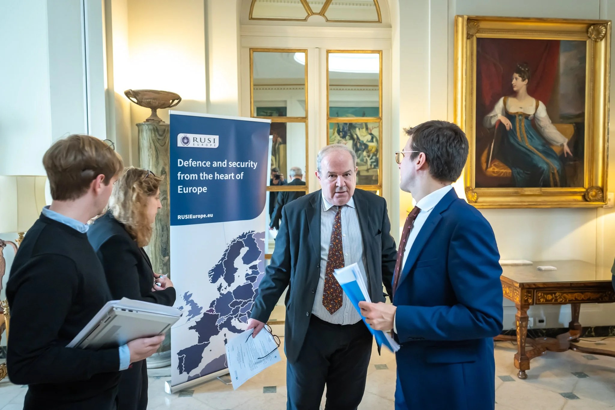 Several people discuss around a "RUSI Europe" banner bearing the text "Defence and security from the heart of Europe" in a room decorated with paintings.