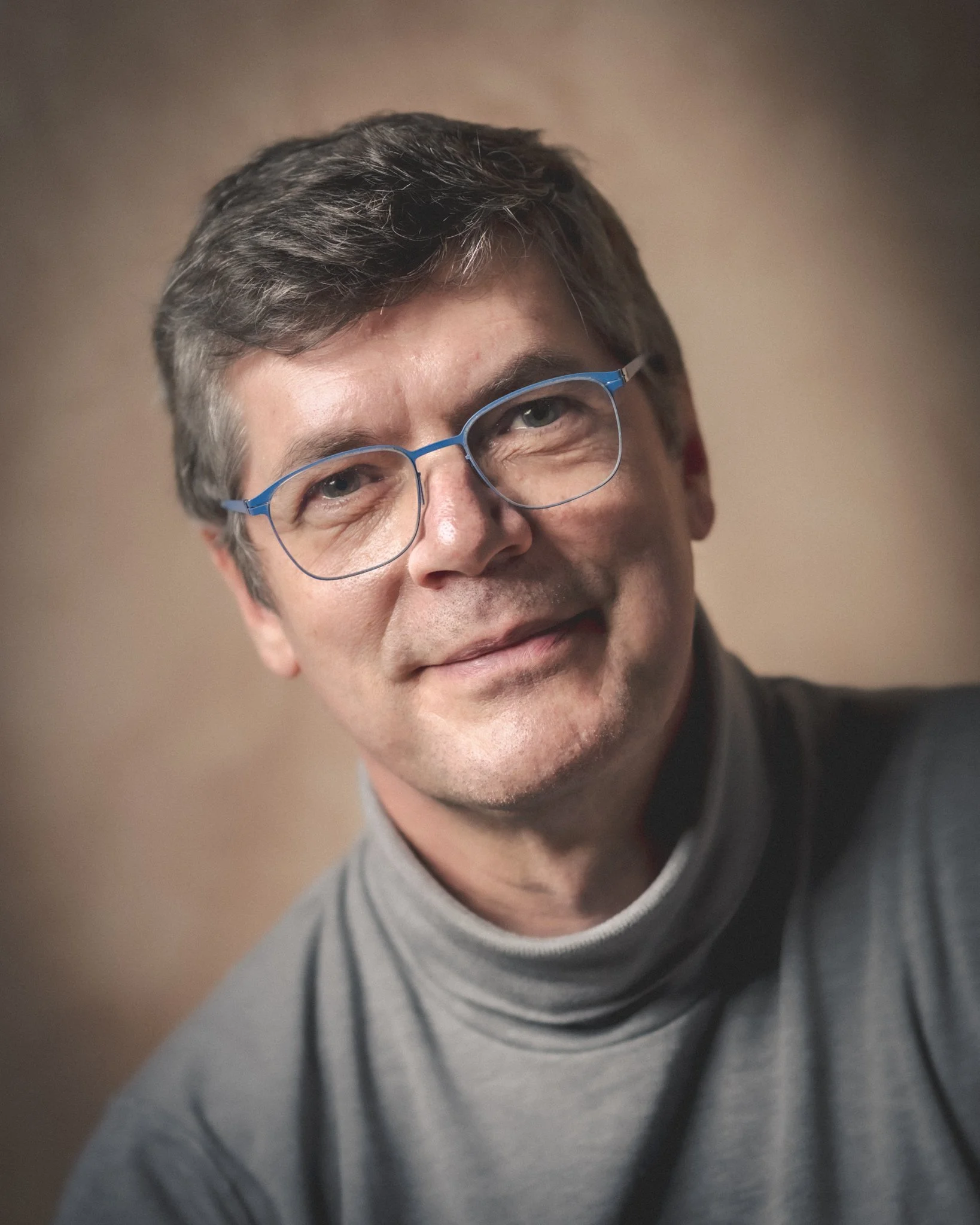 Portrait de Paul Marnef, photographe, portant des lunettes bleues, souriant légèrement, vêtu d'un pull gris, contre un fond neutre.
