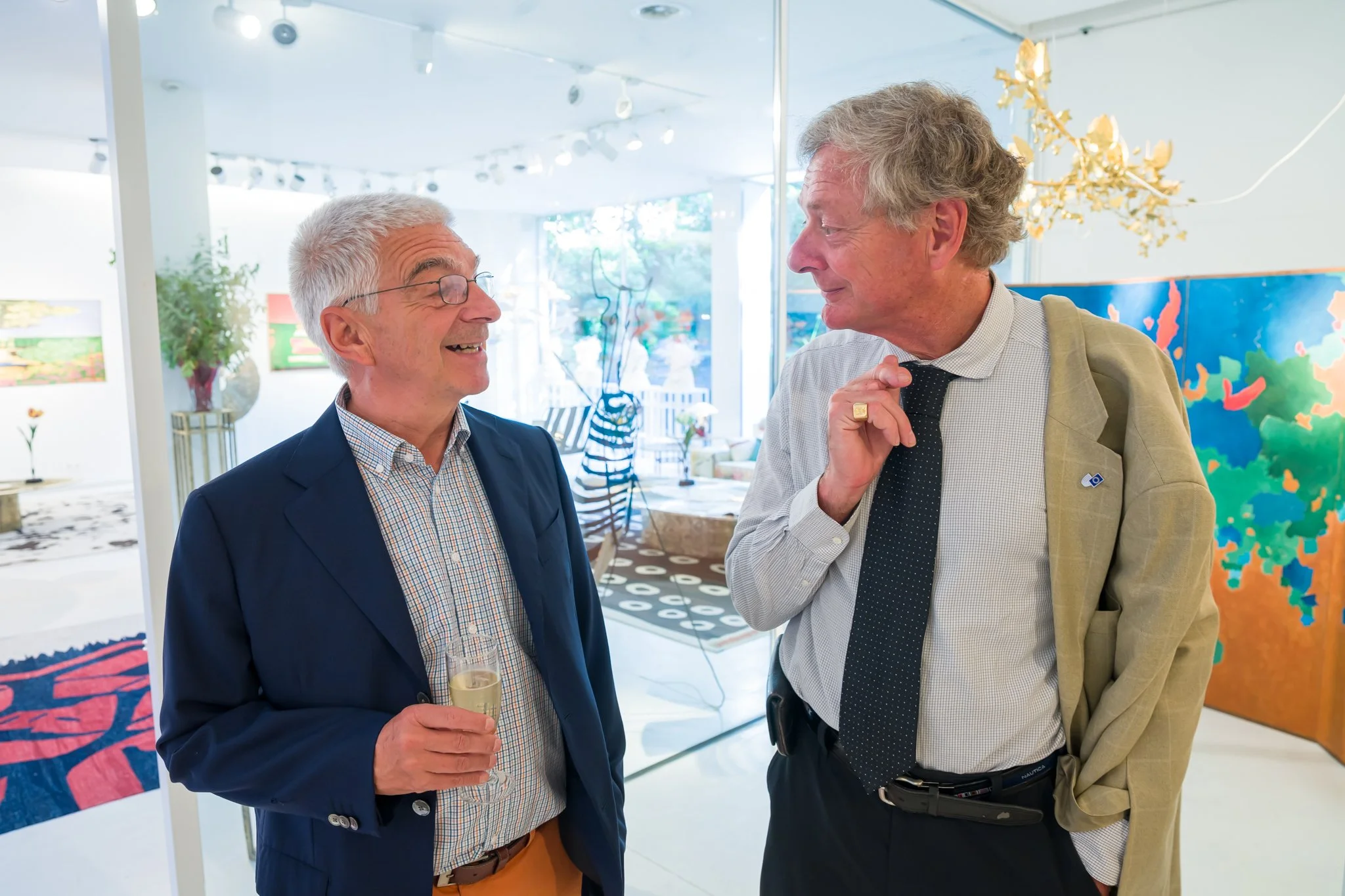 Deux hommes discutent lors d'une exposition dans une galerie d'art, l'un porte un costume bleu avec une robe à carreaux, tenant une coupe de champagne ; l'autre porte un blazer beige, chemise à carreaux et une cravate noire. En arrière-plan, des œuvr