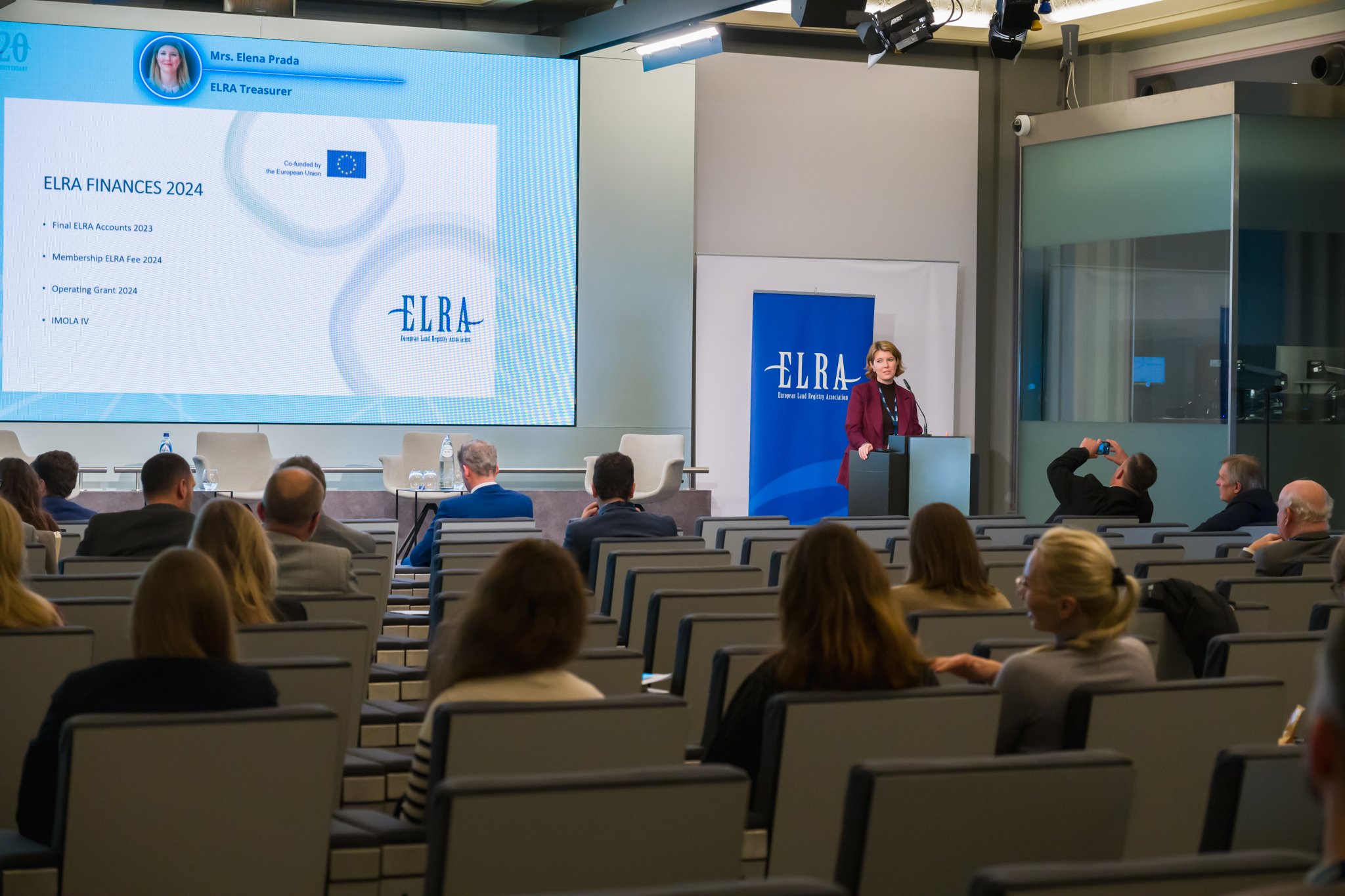 Une conférence sur les finances de l'ELRA 2024 avec une présentatrice debout près d'un pupitre, un écran affichant des informations financières, et un public assis dans une salle de conférence.