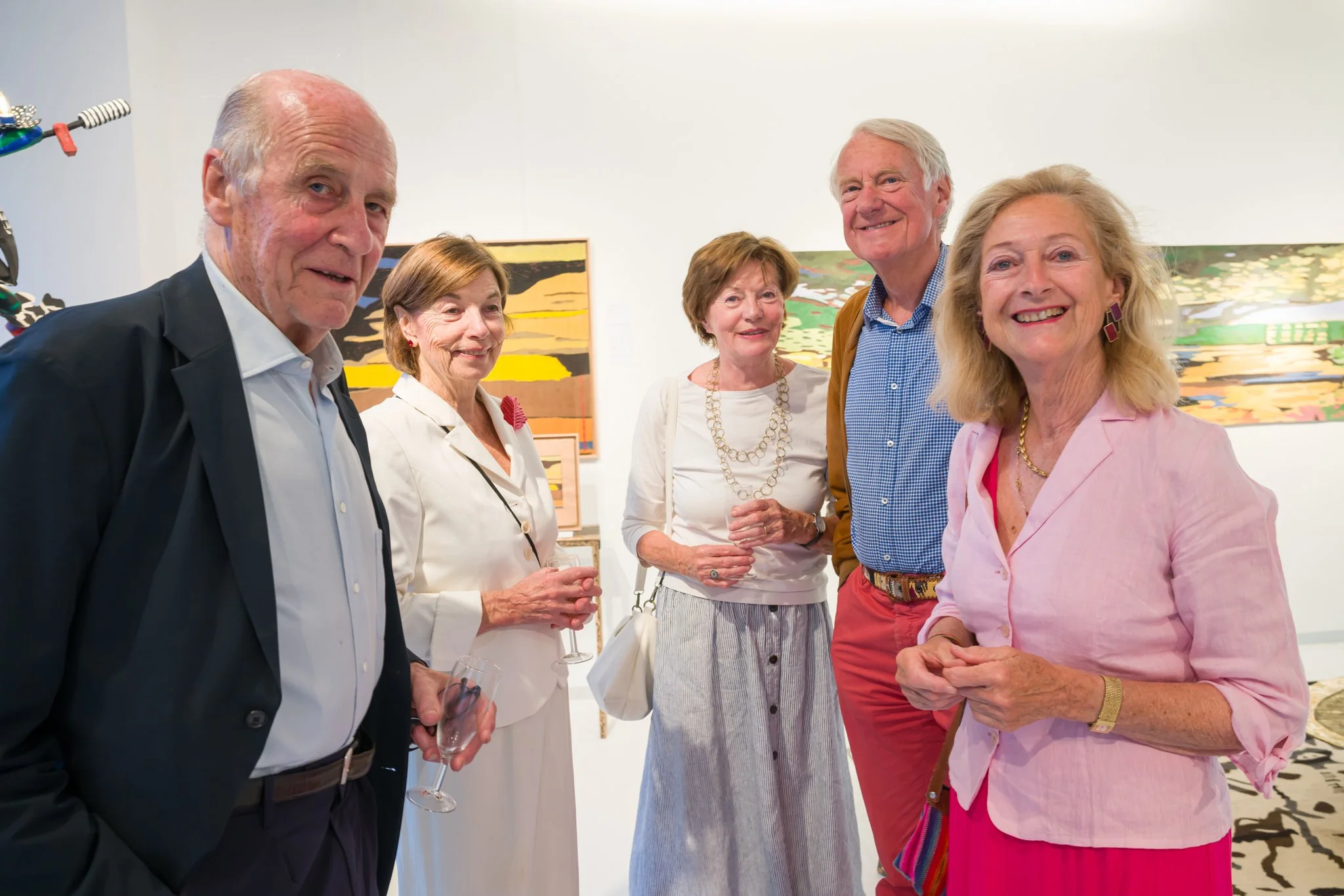 Cinq personnes âgées souriantes lors d'une exposition d'art, posant devant des peintures colorées.