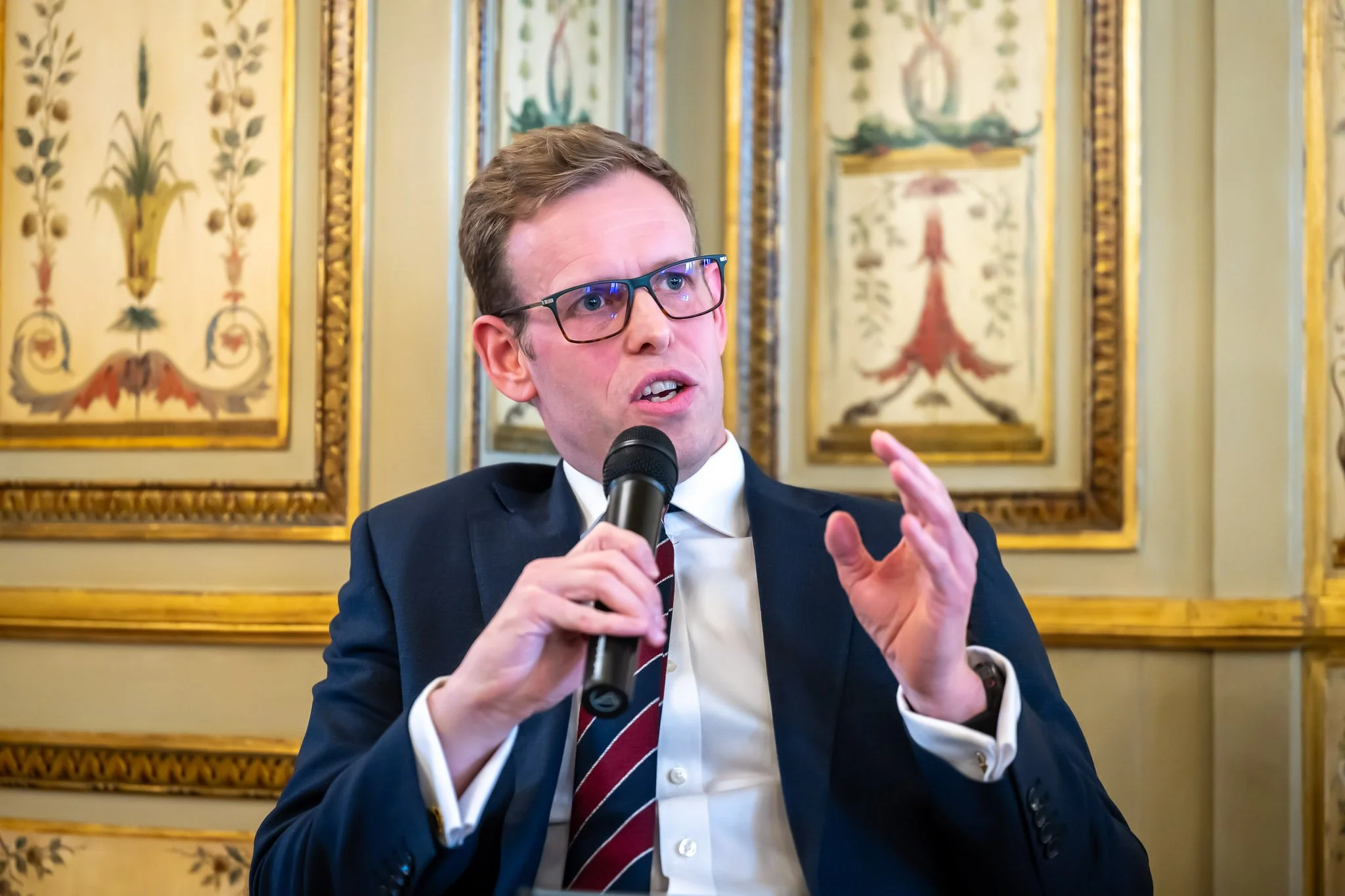A man in a suit speaks into a microphone, gesticulating with his hand, with an ornate wall decoration in the background.