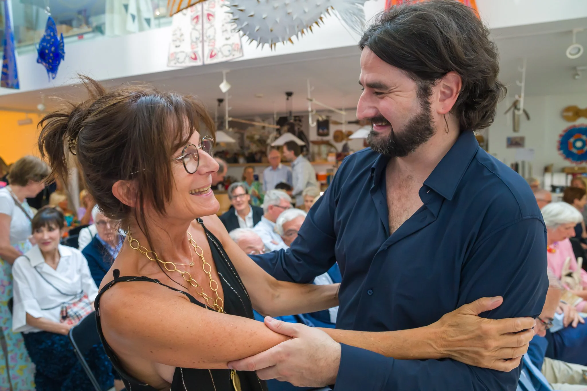 Deux personnes, un homme et une femme, s'embrassent et sourient lors d'une fête ou d'un événement social, avec plusieurs autres personnes en arrière-plan.