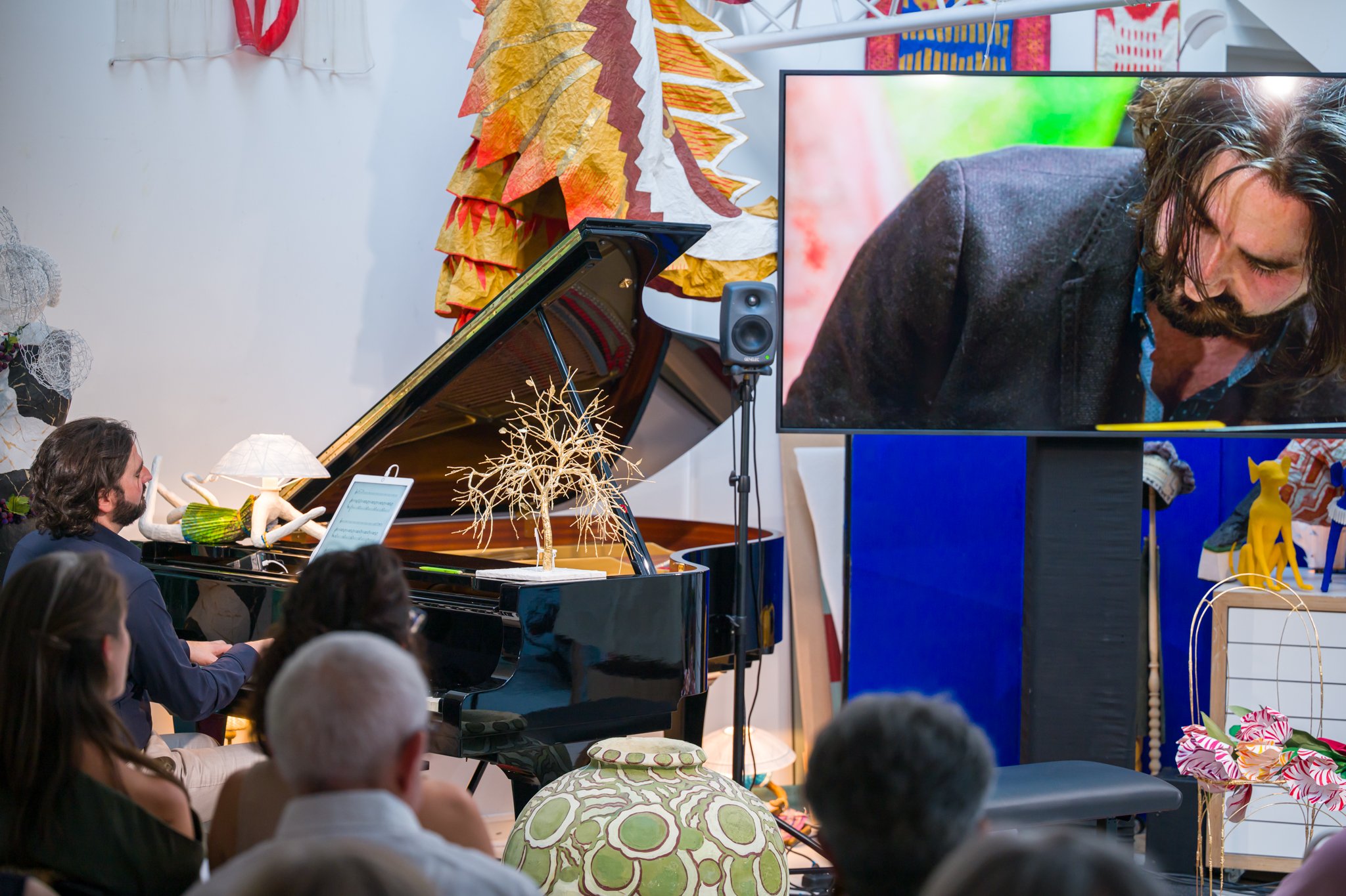 Un groupe de personnes assises regardant un performance musicale avec un piano, un écran vidéo, et des décorations artistiques colorées dans une salle d'exposition.