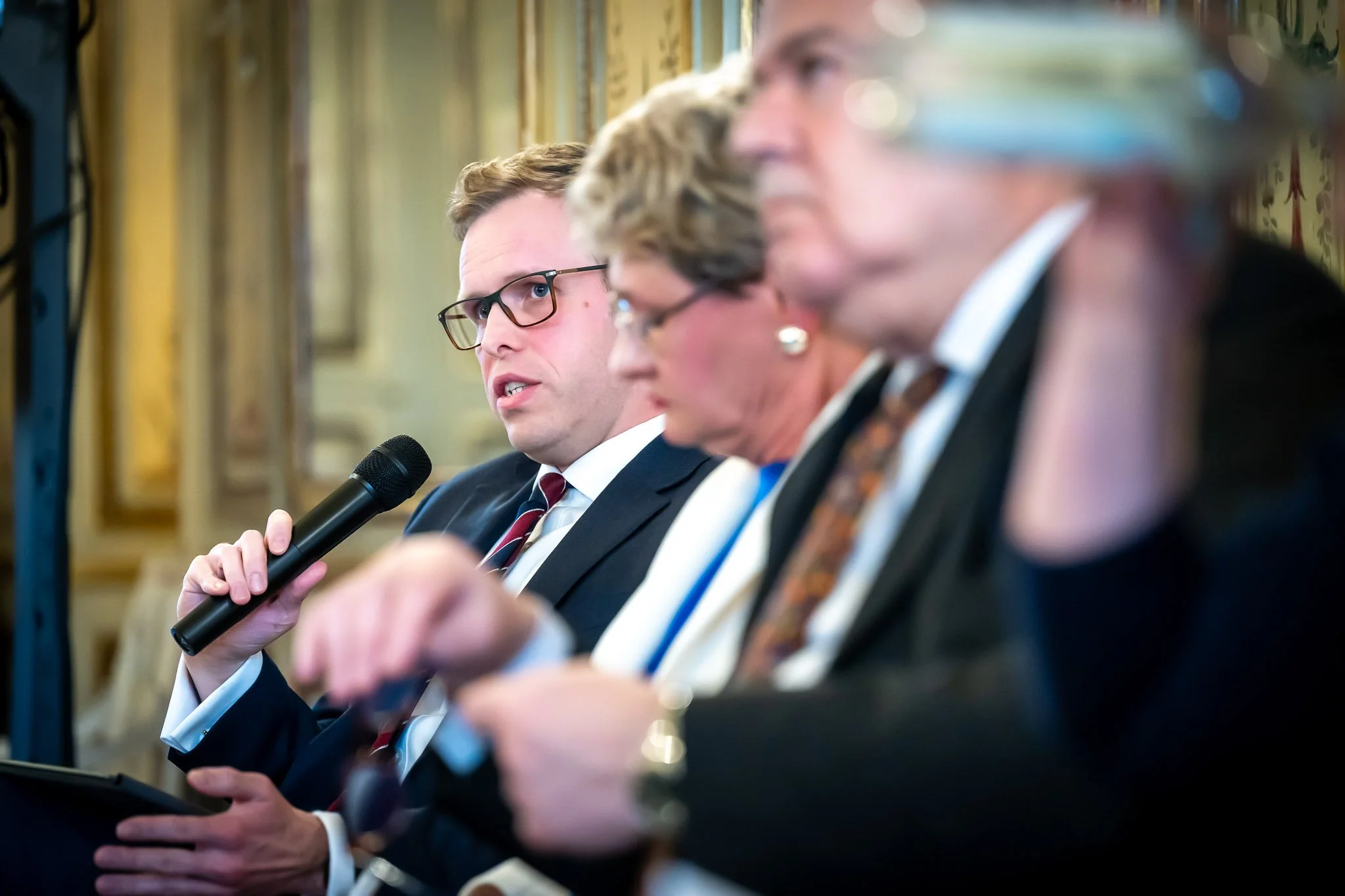 Three people in suits chatting at an event, one man holding a microphone.