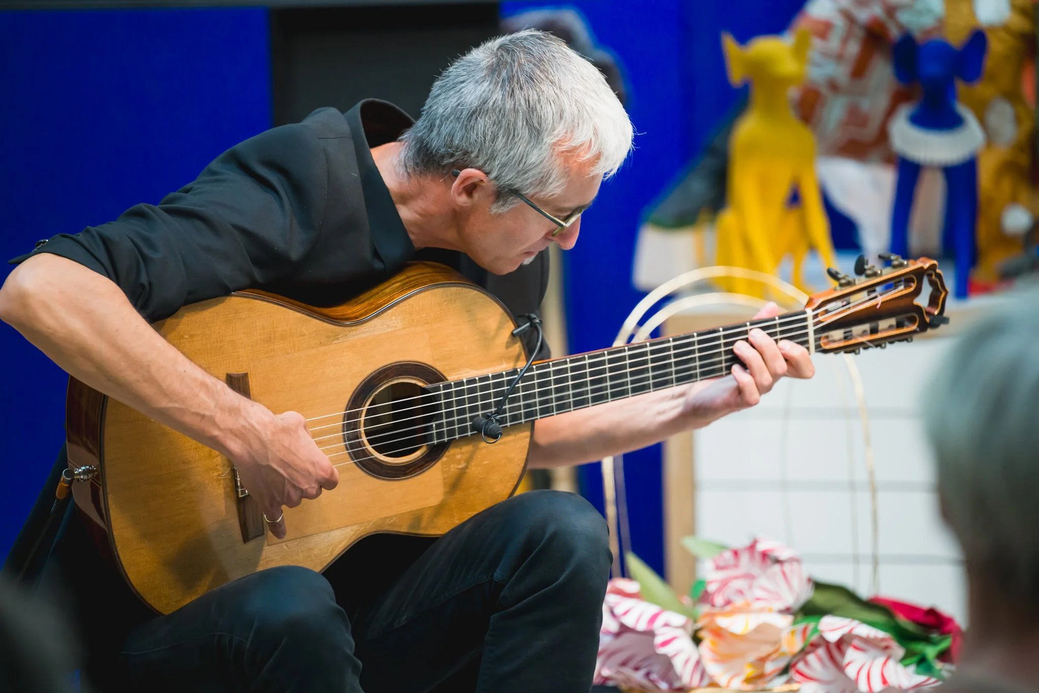 Un homme avec des cheveux gris joue de la guitare acoustique lors d'une performance, assis devant un fond coloré.