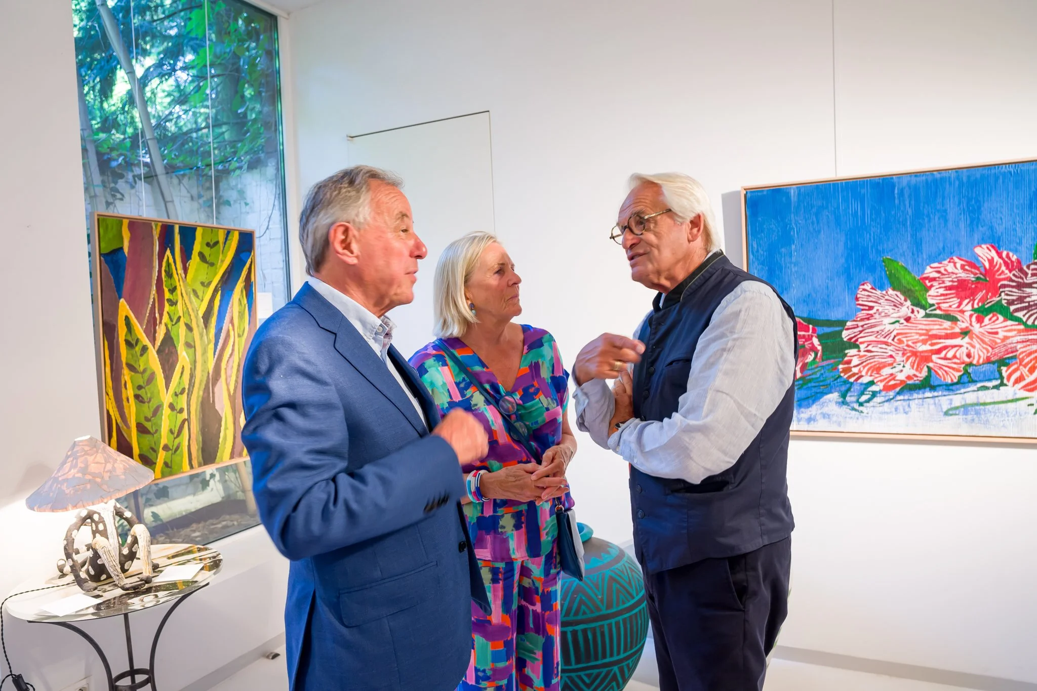 Trois personnes discutent dans une galerie d'art avec des œuvres colorées en arrière-plan.
