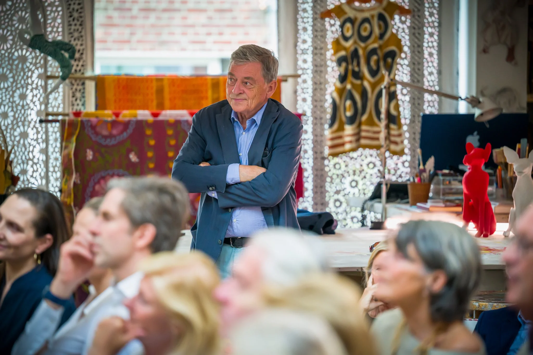 Un homme âgé en blazer bleu et chemise bleue, avec des bras croisés, regarde à gauche lors d'une réunion ou conférence, entouré par plusieurs personnes. La salle est décorée avec des tissus colorés et des objets artistiques.