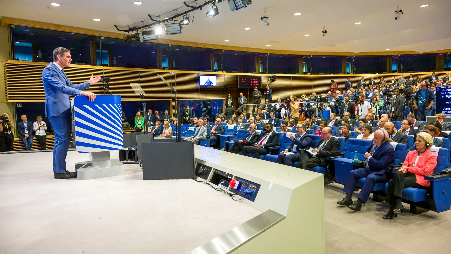 Un homme en costume bleu donne un discours sur un podium devant un public dans une salle de conférence moderne. Des écrans et caméras sont visibles, ainsi que des personnes prenant des notes et portant des casques audio.