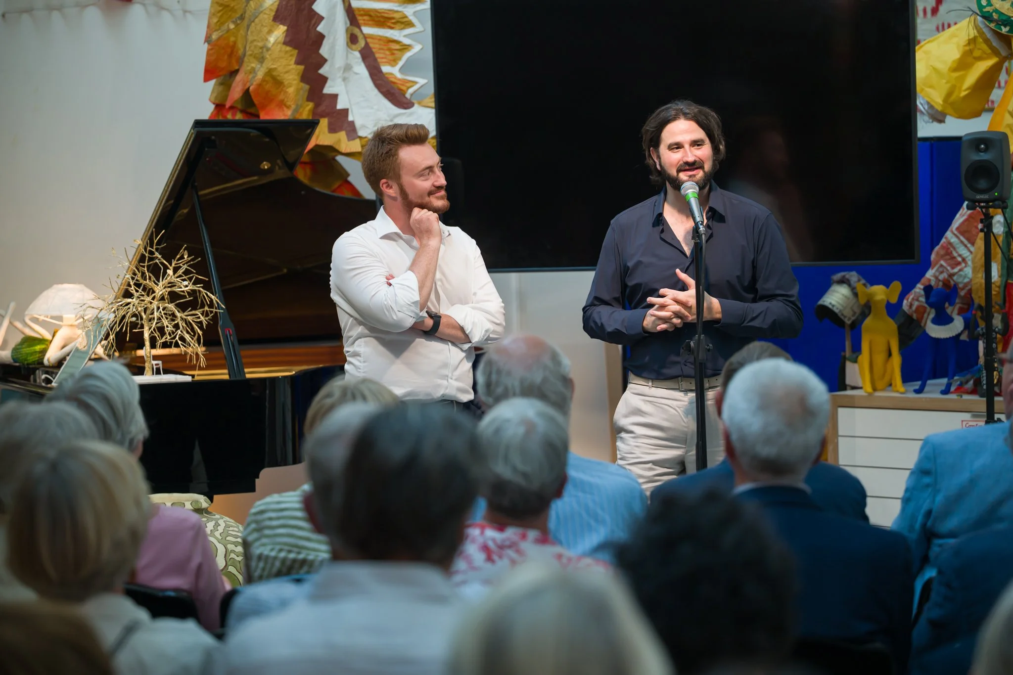 Deux hommes parlent lors d'une présentation ou d'un événement avec une audience. Un homme parle dans un microphone, l'autre écoute attentivement. En arrière-plan, un piano et des décorations colorées.