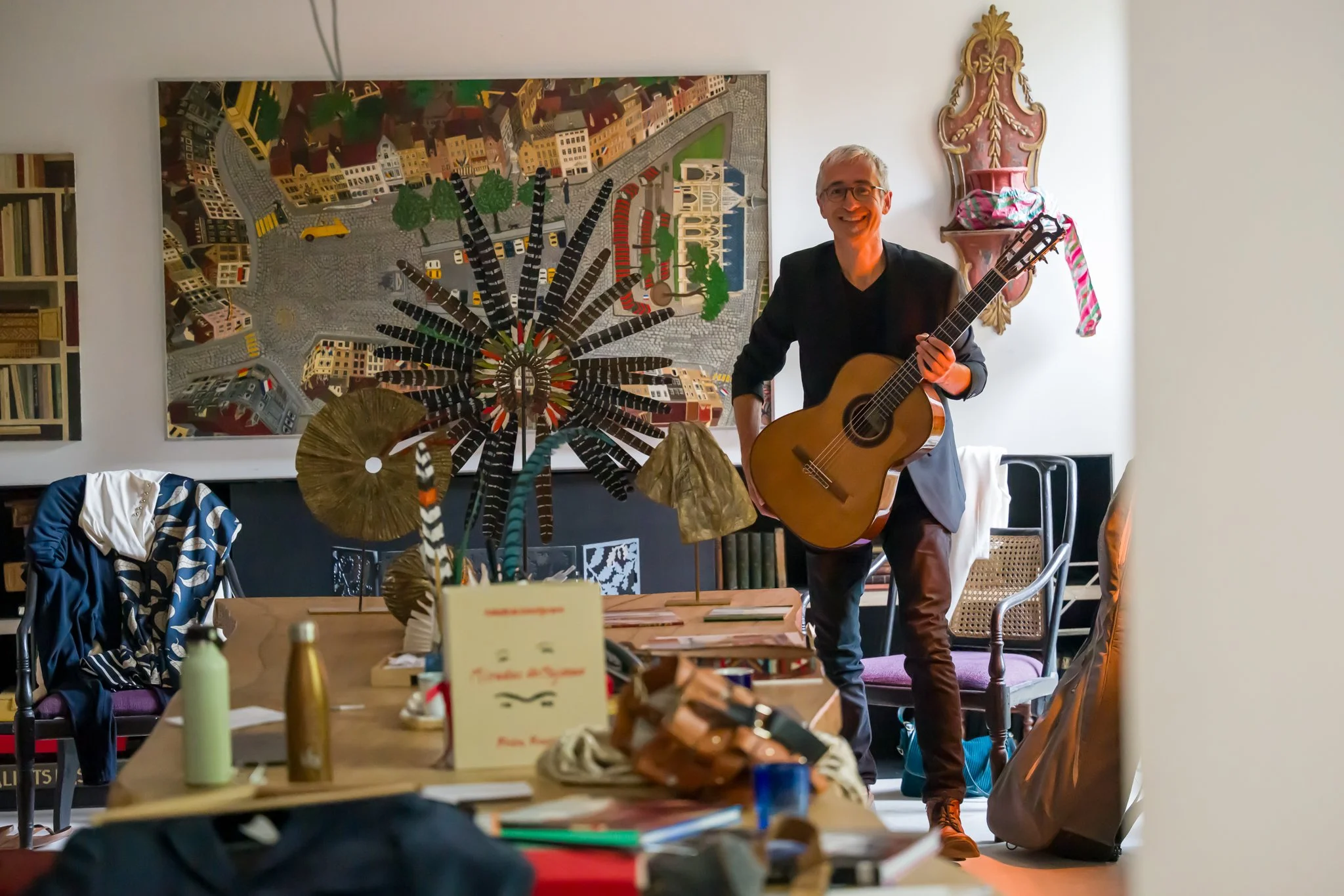 Un homme souriant jouant de la guitare dans une pièce décorée avec un tableau coloré et diverses décorations.