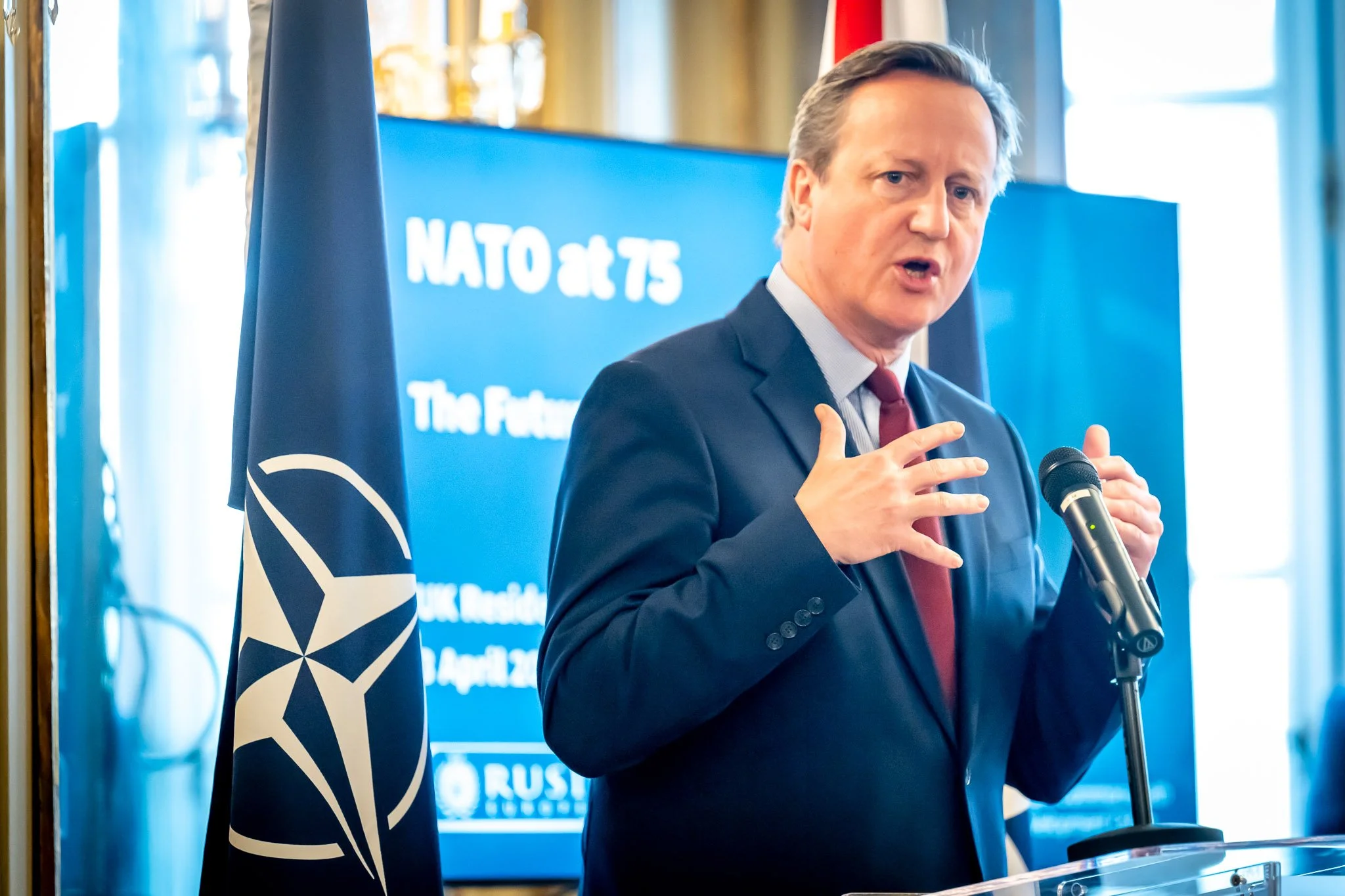 A man in a suit speaks into a microphone at an event celebrating 75 years of NATO, with a NATO flag beside him.