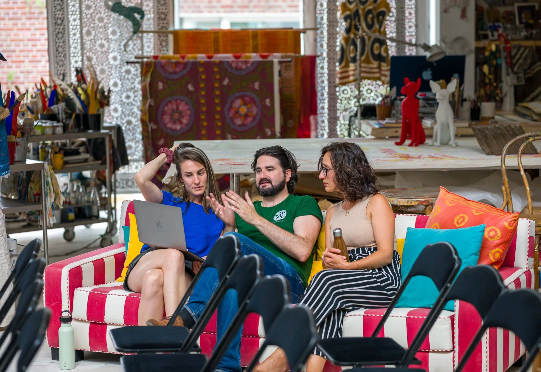 Trois personnes discutent sur un canapé coloré dans un atelier créatif, avec des chaises noires devant eux et des objets artistiques en arrière-plan.