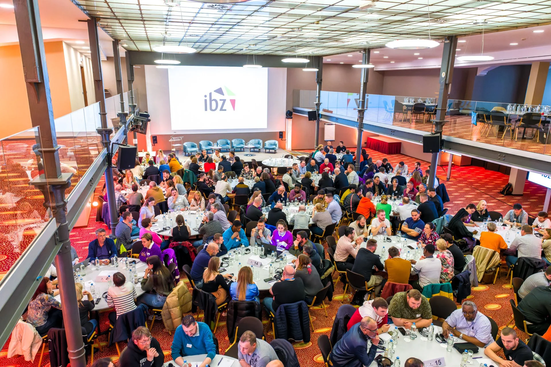 Salle de conférence avec de nombreuses personnes assises autour de tables rondes, projetant un écran affichant le logo "ibz".