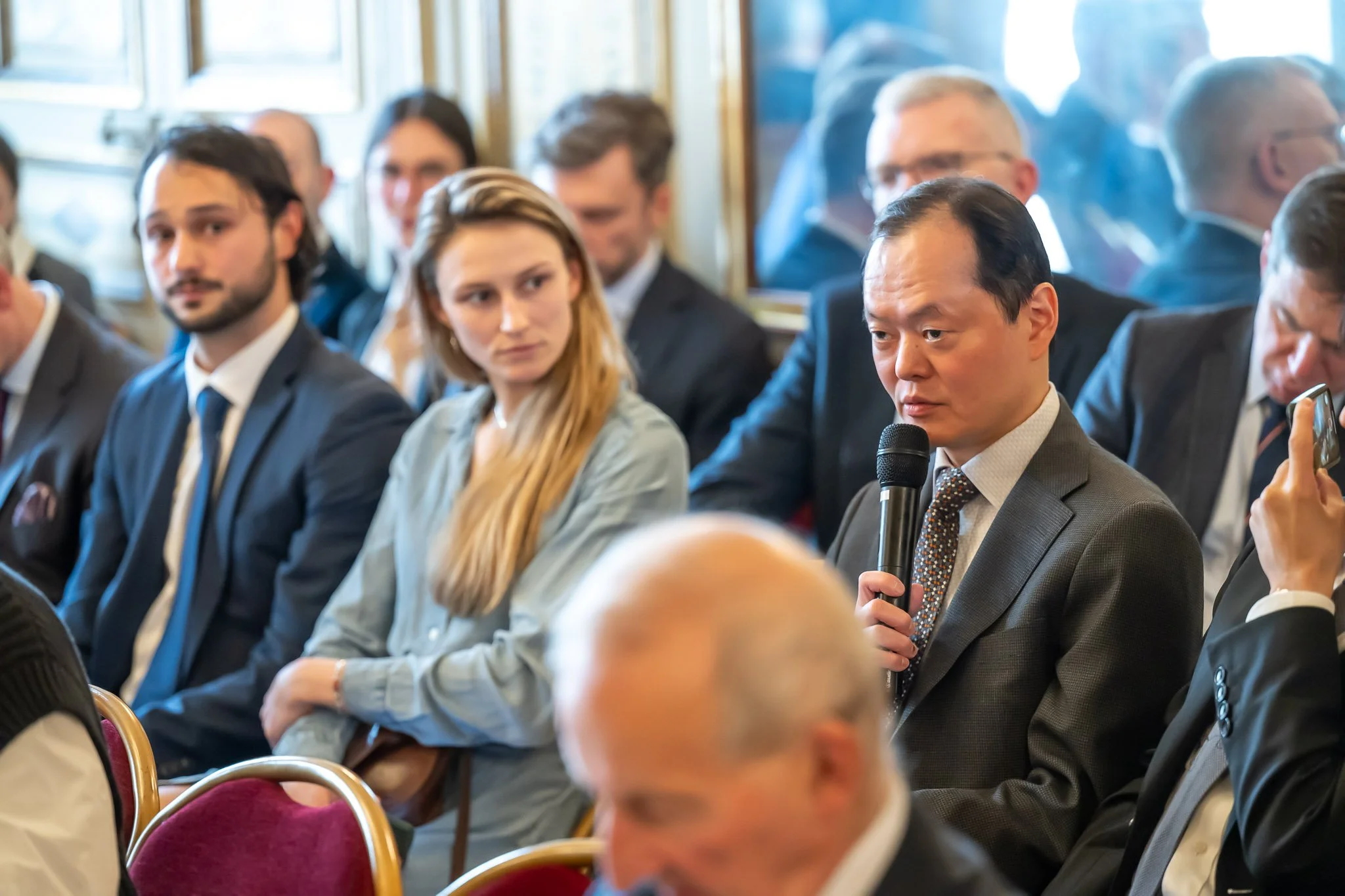 A group of people in suits seated at a conference, with a man holding a microphone.