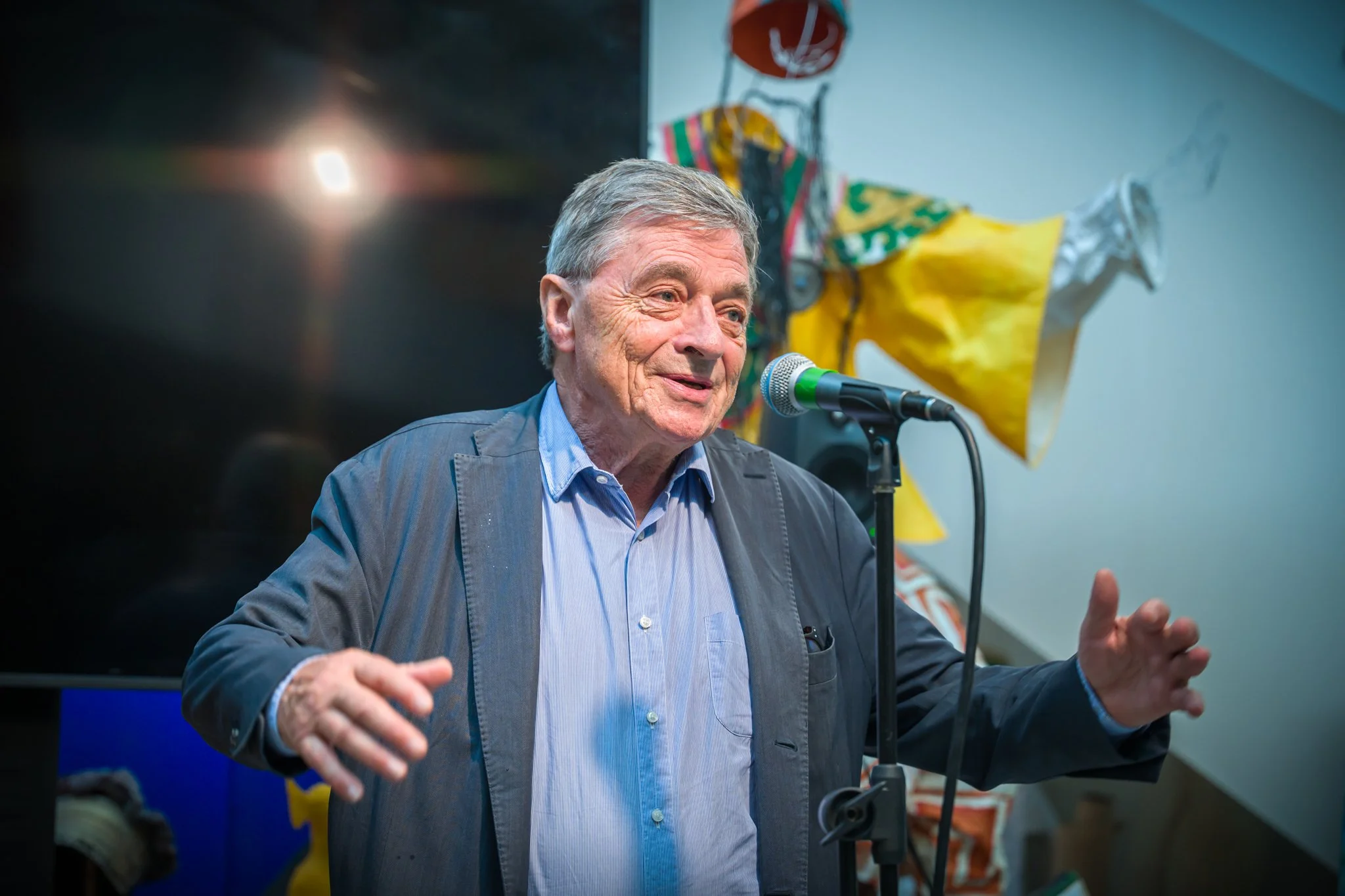 Un homme âgé parle dans un microphone, vêtements décontractés, fond coloré avec des drapeaux ou décorations artisanales.