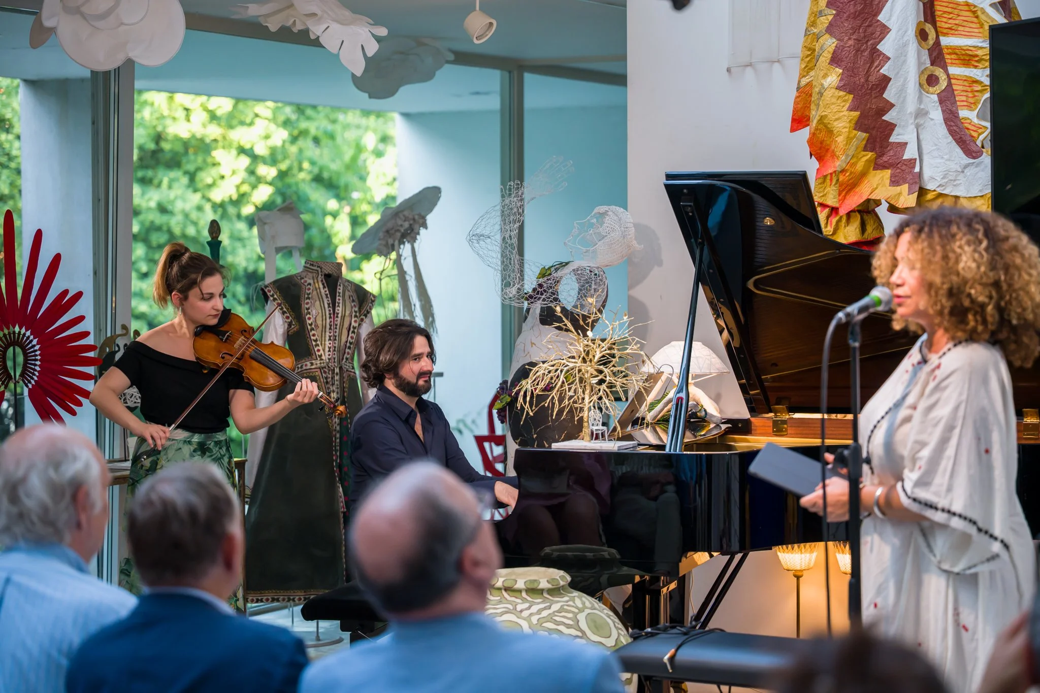Un groupe de musiciens jouant lors d'une performance dans un intérieur décoré avec des œuvres d'art, un piano, une femme chantant au micro, et un public en fauteuils. La scène est décorée avec des éléments colorés et des décorations artistiques, avec
