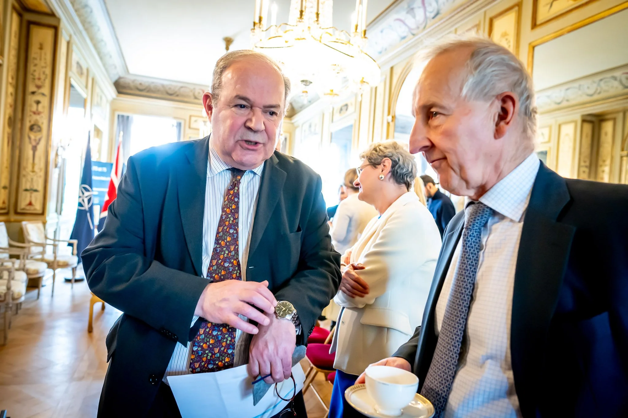 Two men in suits are chatting in an elegant room, with people in the background. One is holding a cup of coffee and the other is looking at his watch. In the background, a NATO flag is visible.