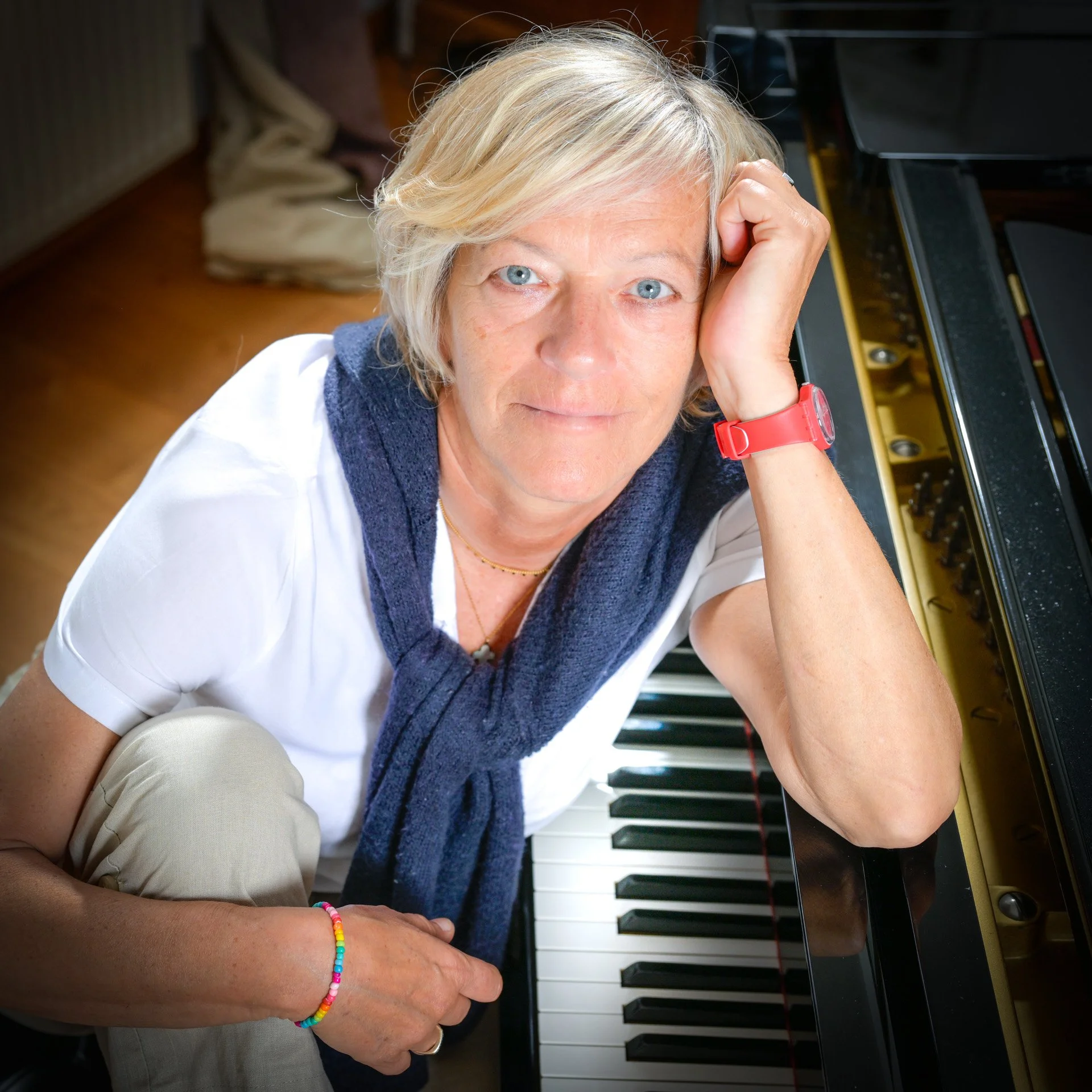 Fabienne Vignier, une femme avec des cheveux blonds, portant une chemise blanche, avec un pull bleu autour du cou, assise à un piano et regardant l'objectif.