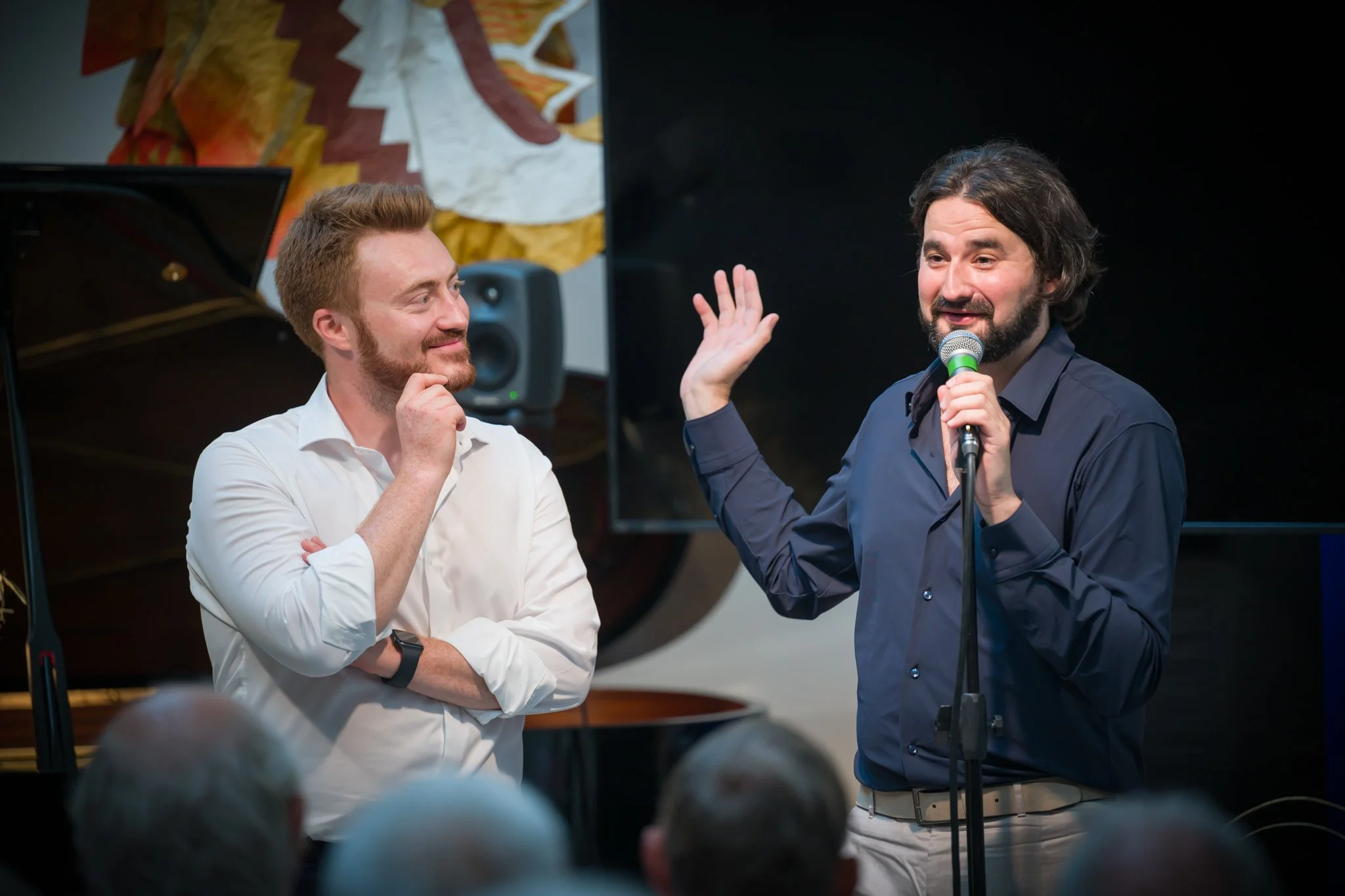 Deux hommes discutent sur scène lors d’une performance ou d’un événement. L’un parle au micro, l’autre semble écouter attentivement, dans un environnement intérieur avec un piano en arrière-plan.