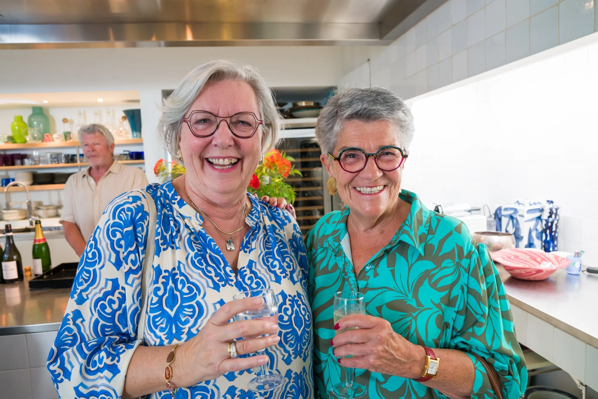 Deux femmes âgées souriantes tenant des verres de vin dans une cuisine, célébrant un événement.