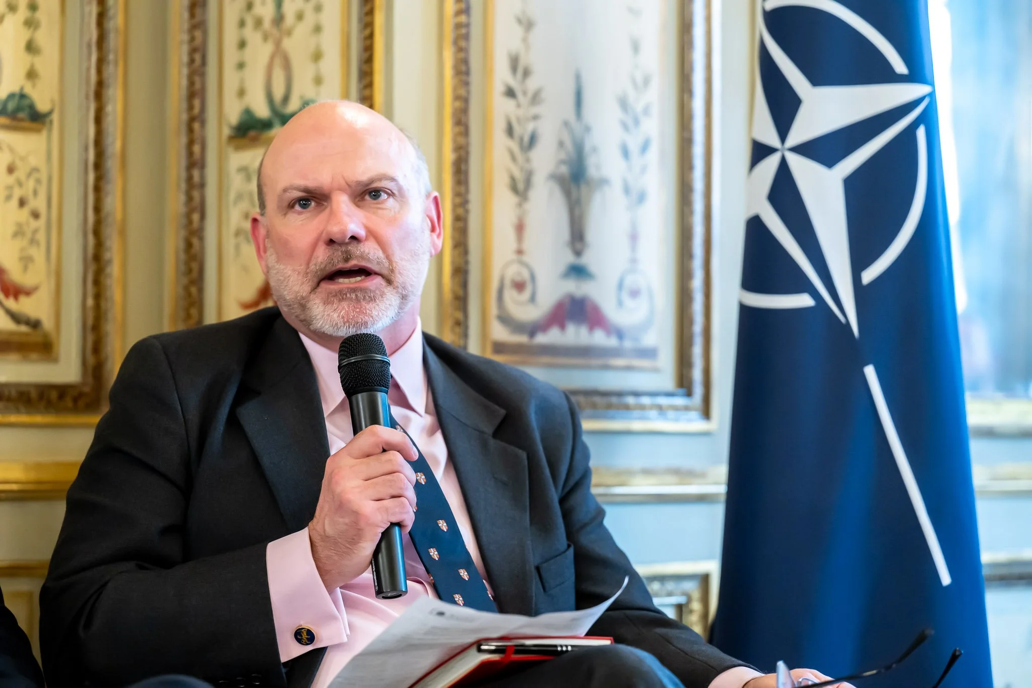 Man in suit speaking into a microphone with a NATO flag beside him.