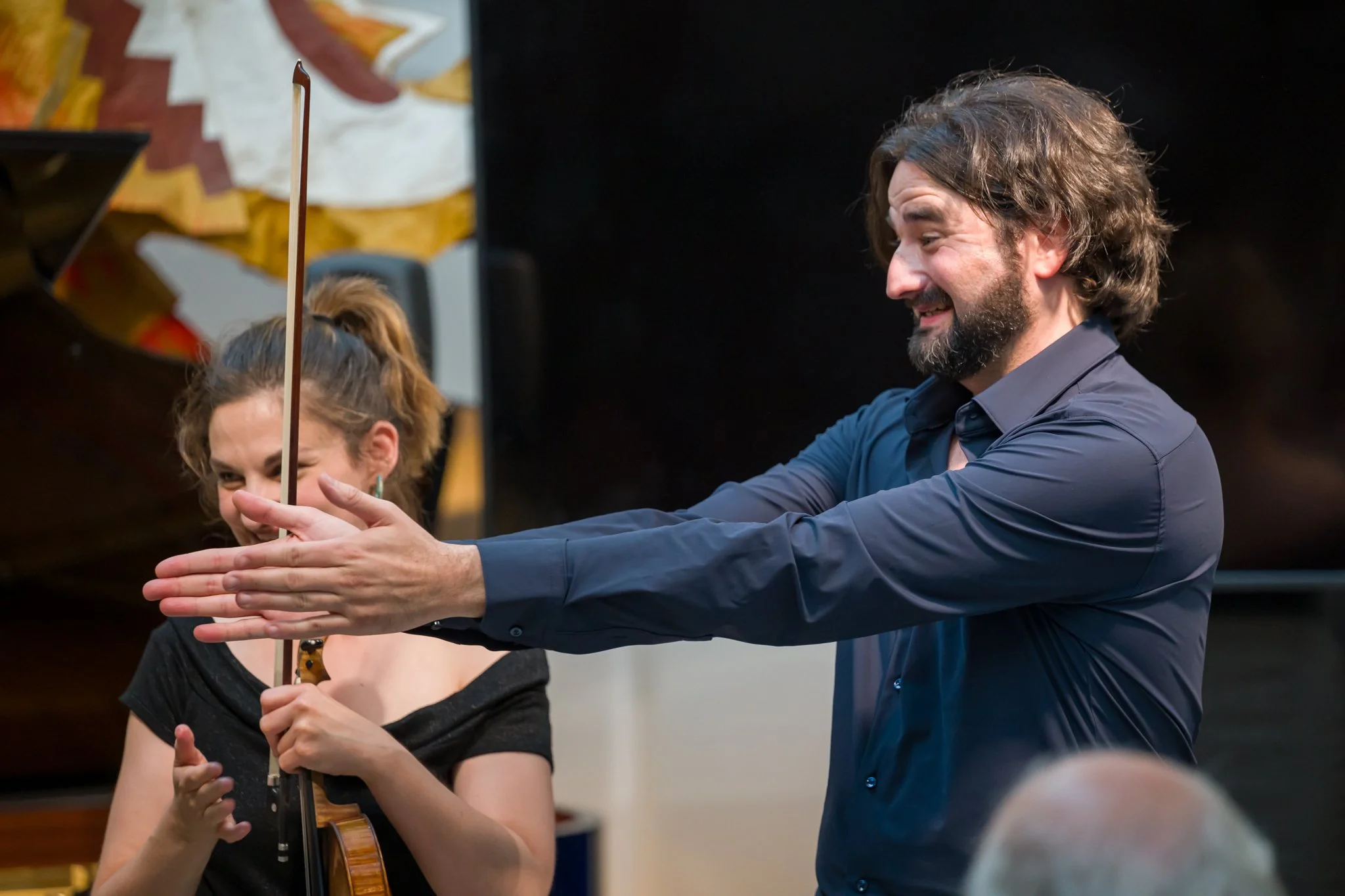 Un homme et une femme, probablement dans un contexte musical, l'homme semble diriger ou enseigner, tandis que la femme joue du violon dans une salle de musique.