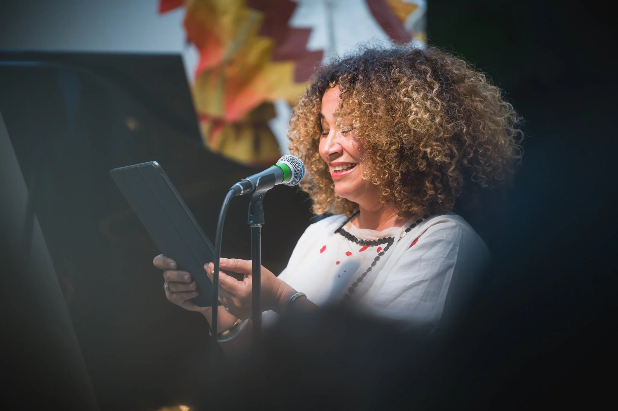 Femme avec des cheveux bouclés, souriante, lisant ou parlant dans un microphone lors d'une performance ou d'une présentation, avec un fond coloré et un piano en arrière-plan.