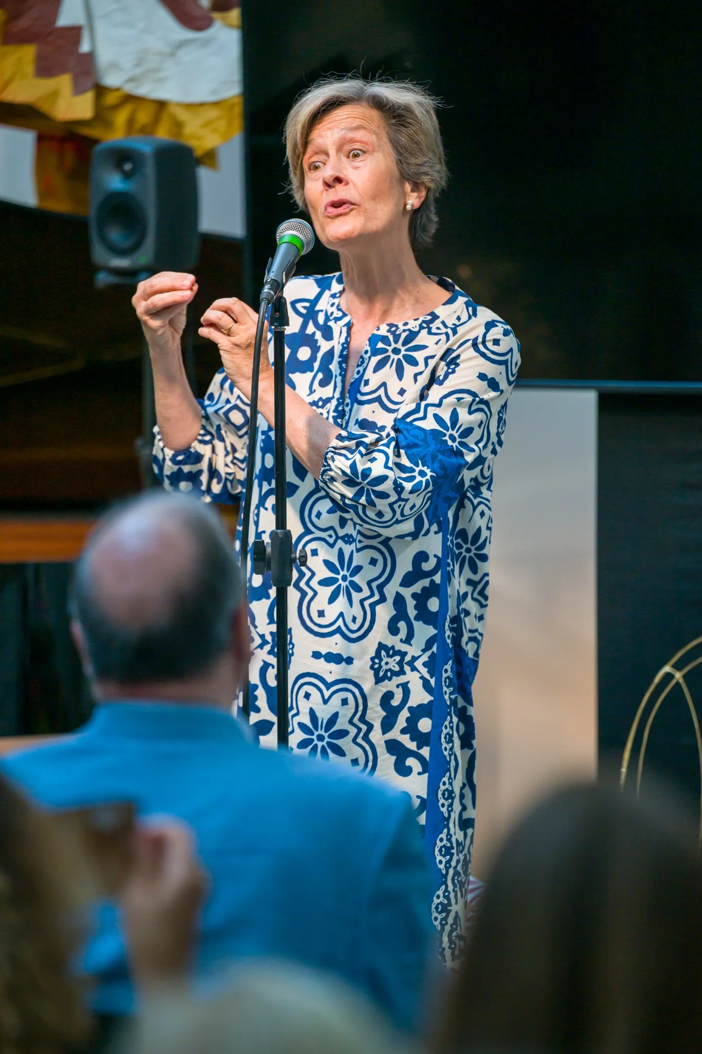 Femme âgée parlant lors d'une présentation ou d'une conférence, portant une robe à motifs bleus et blancs, devant un microphone, en présence d'un auditoire.