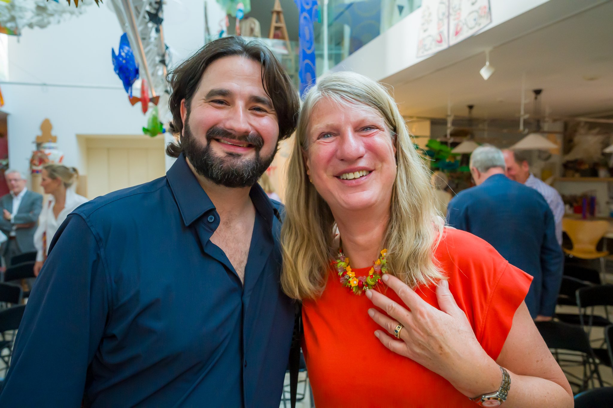 Deux personnes souriantes, un homme avec une barbe et une femme blonde, posant ensemble lors d'un événement social en intérieur, avec d'autres personnes en arrière-plan.