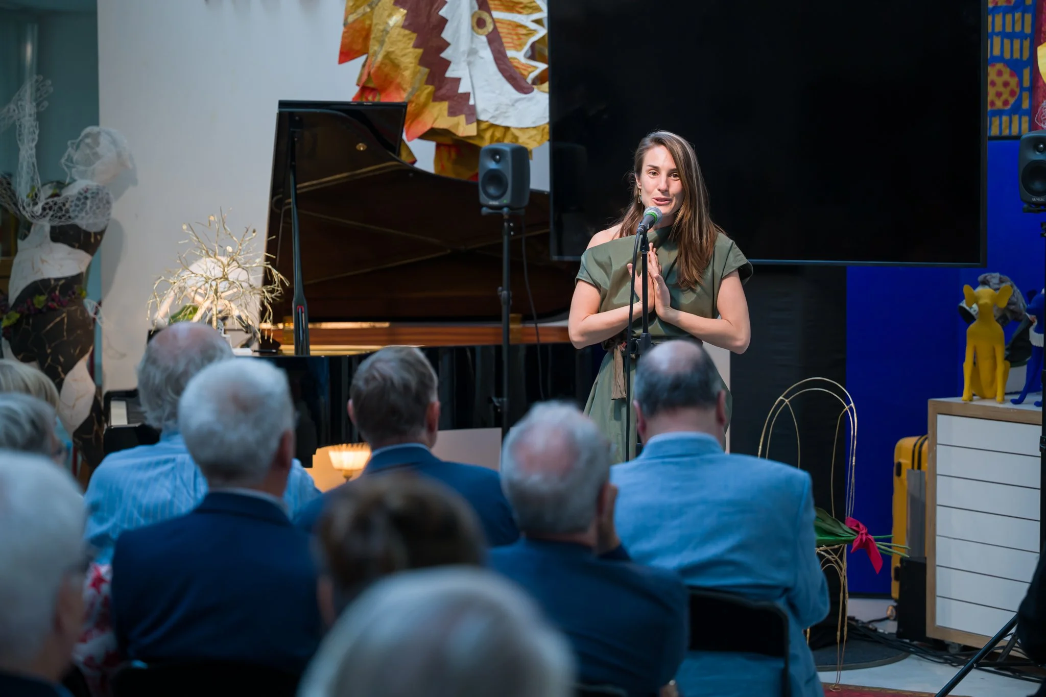 Une femme parle devant un public lors d'une performance ou d'une présentation, une grande télévision à l'arrière, un piano, et des sculptures colorées dans la salle.