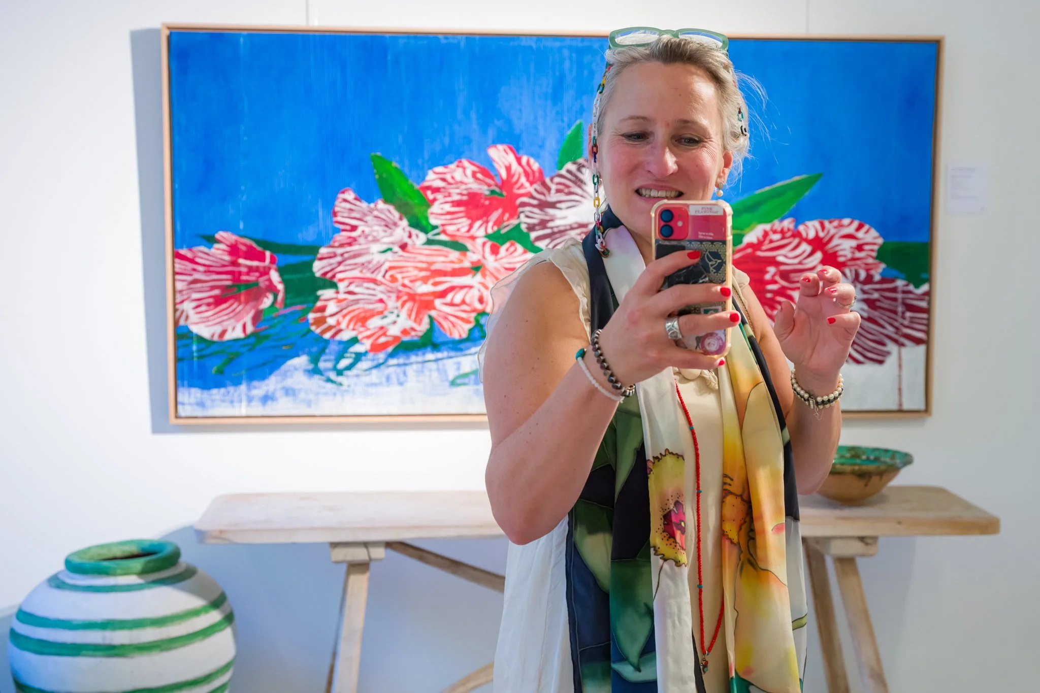 Une femme souriante prenant une autoportrait dans une galerie d'art avec un tableau coloré de fleurs en arrière-plan.