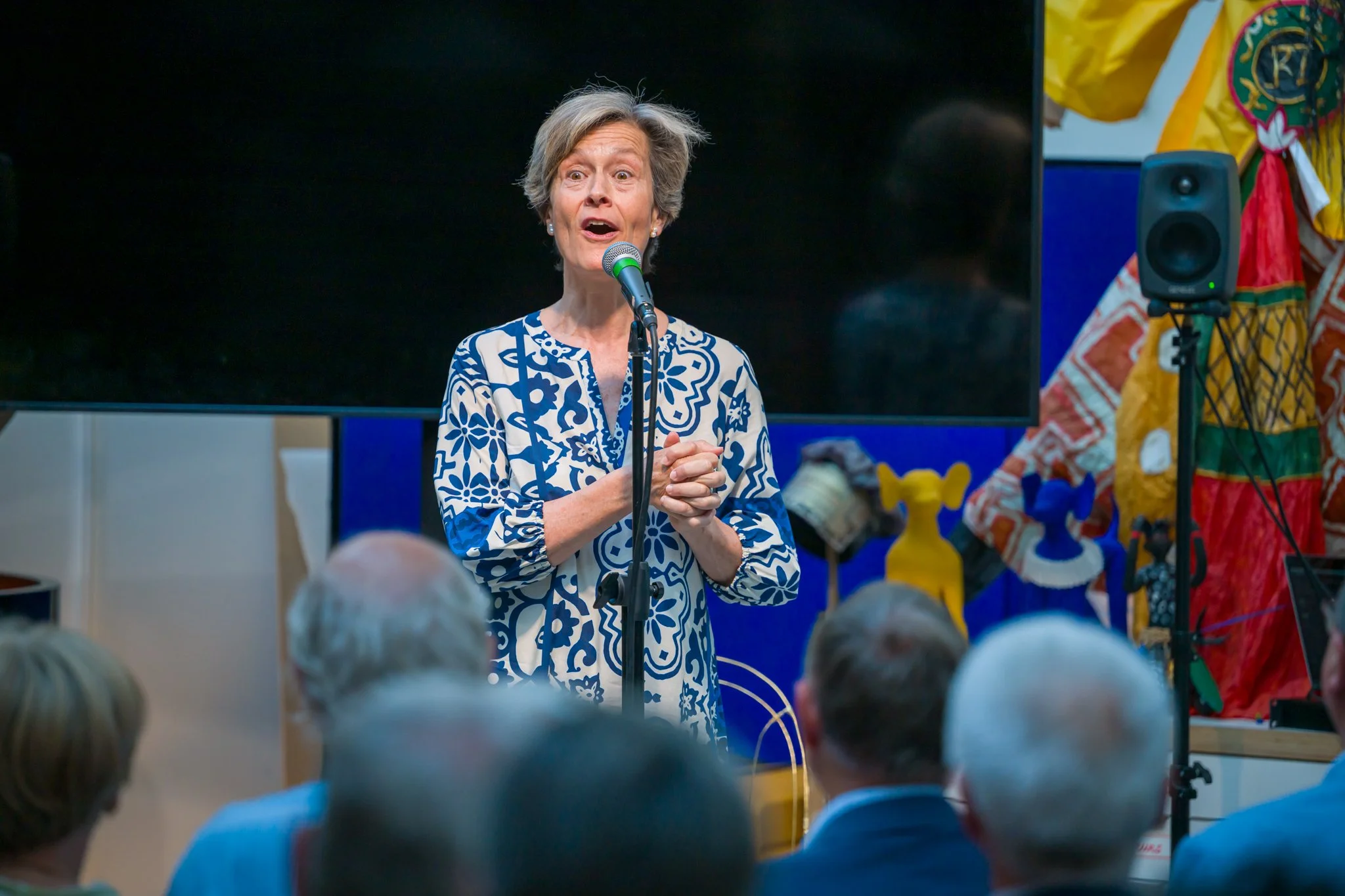 Une femme âgée avec des cheveux courts gris, portant une chemise à motifs bleus et blancs, parle ou chante devant un microphone lors d'une présentation ou d'un événement, avec un groupe de personnes debout devant elle, dans un espace intérieur.